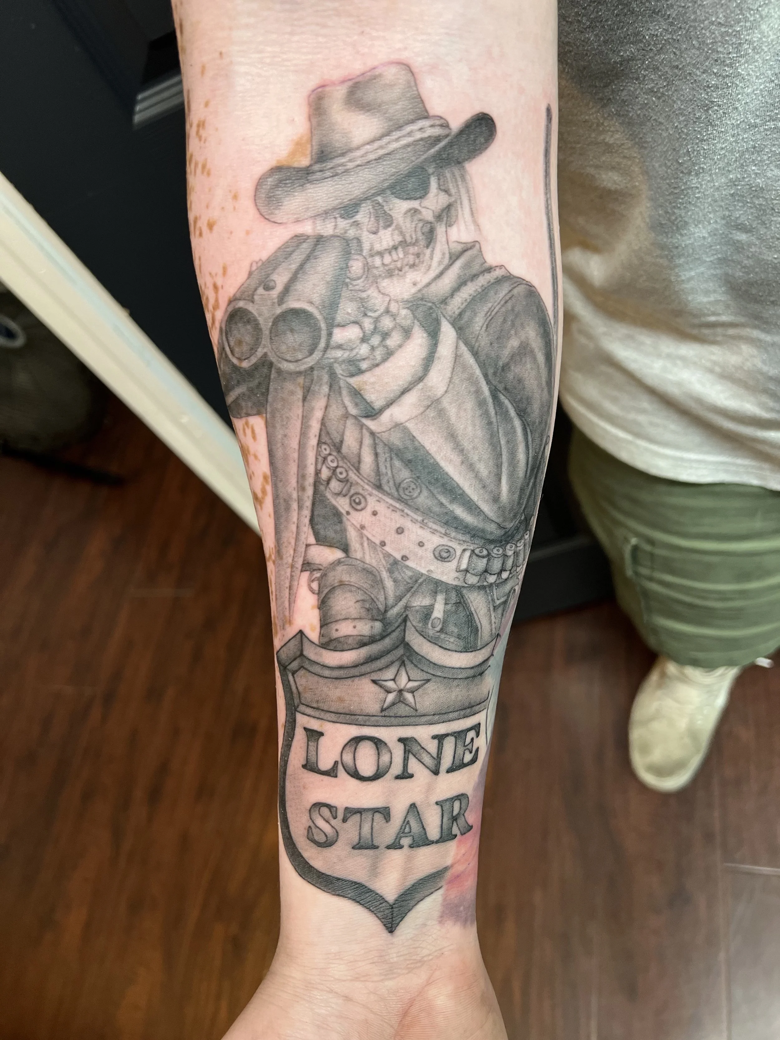 A black and gray tattoo of a skeleton sheriff wearing a cowboy hat, holding two revolvers, with a sheriff star badge below and the words "LONE STAR".