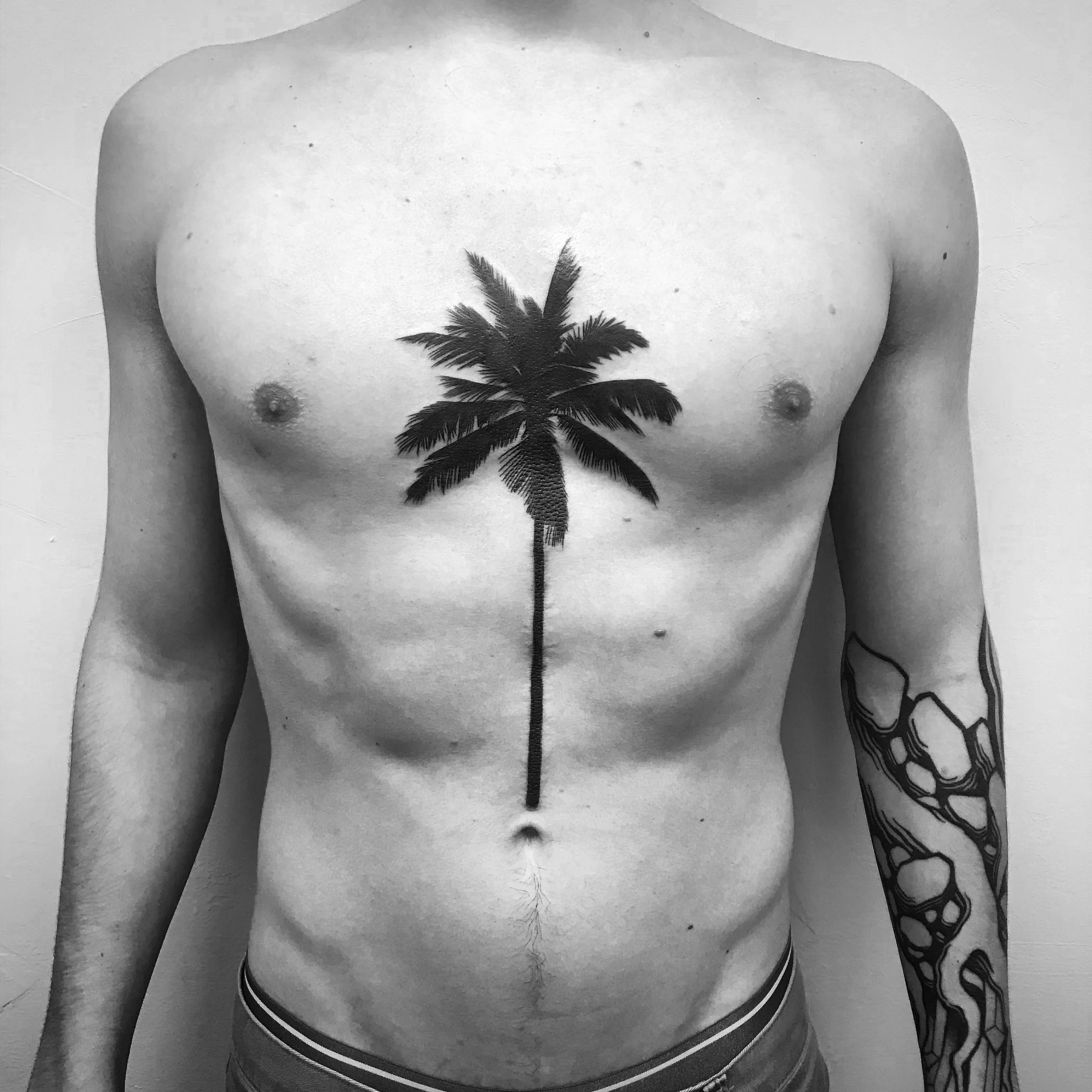 Male torso with a palm tree tattoo on the chest and a sleeve tattoo on the right arm in black and white.