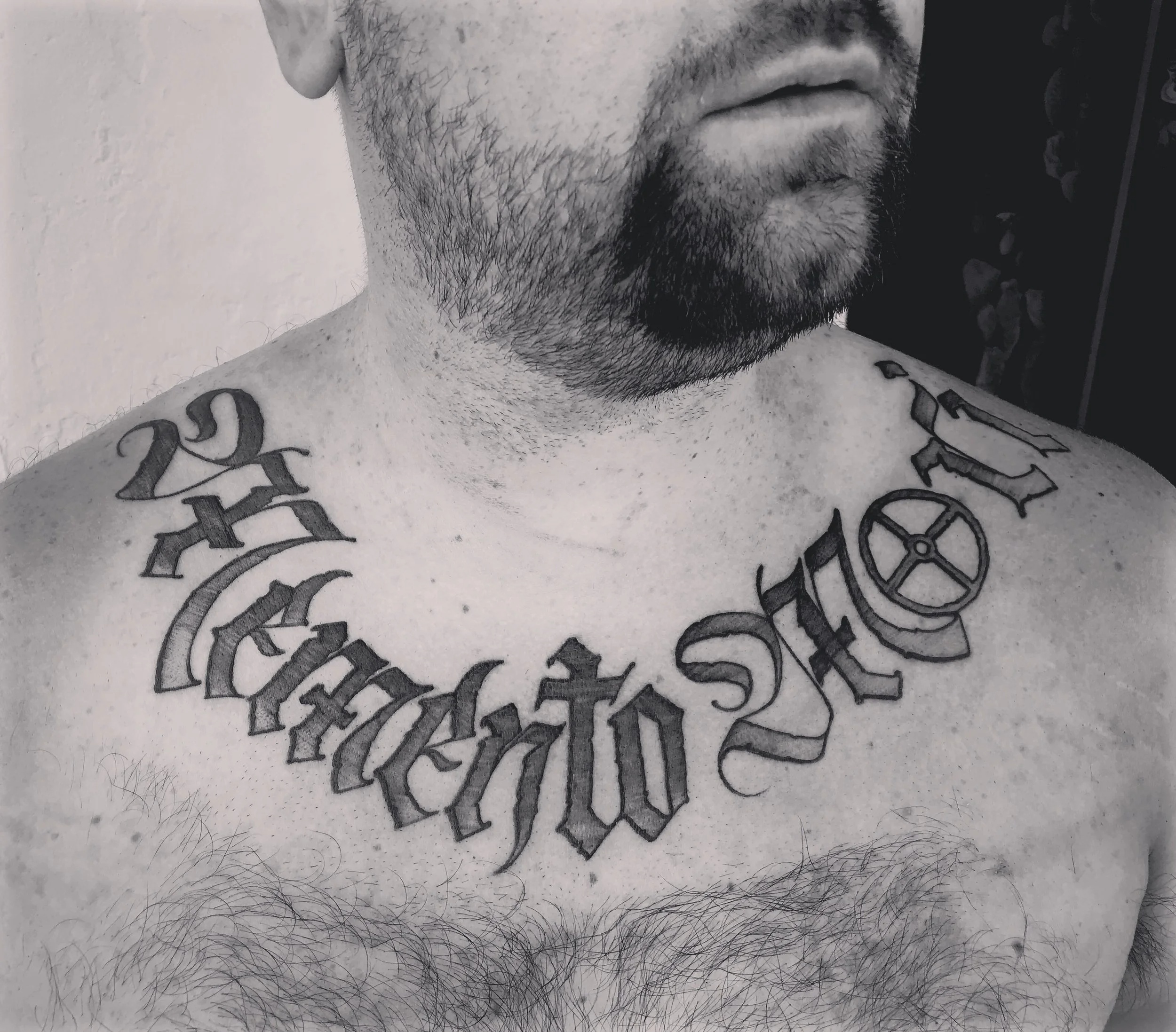 Close-up of a man's chest and neck, showing a tattoo with the words 'memento mori'  in ornate gothic script across the collarbone and a heart-shaped lock on the right side.