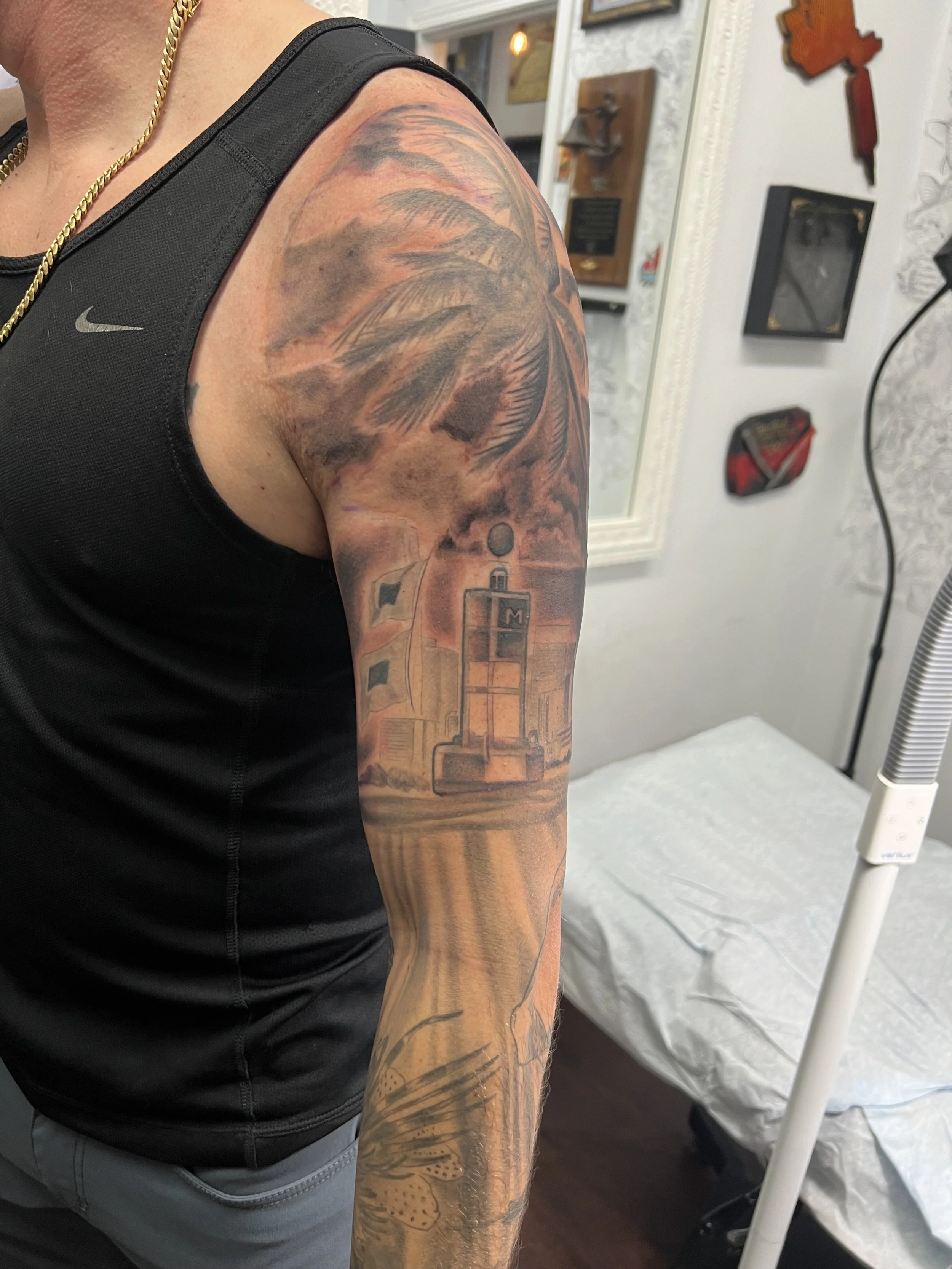 Tattoo of a lighthouse, flags, and clouds on a person's upper arm and shoulder, with a palm tree and a butterfly below.