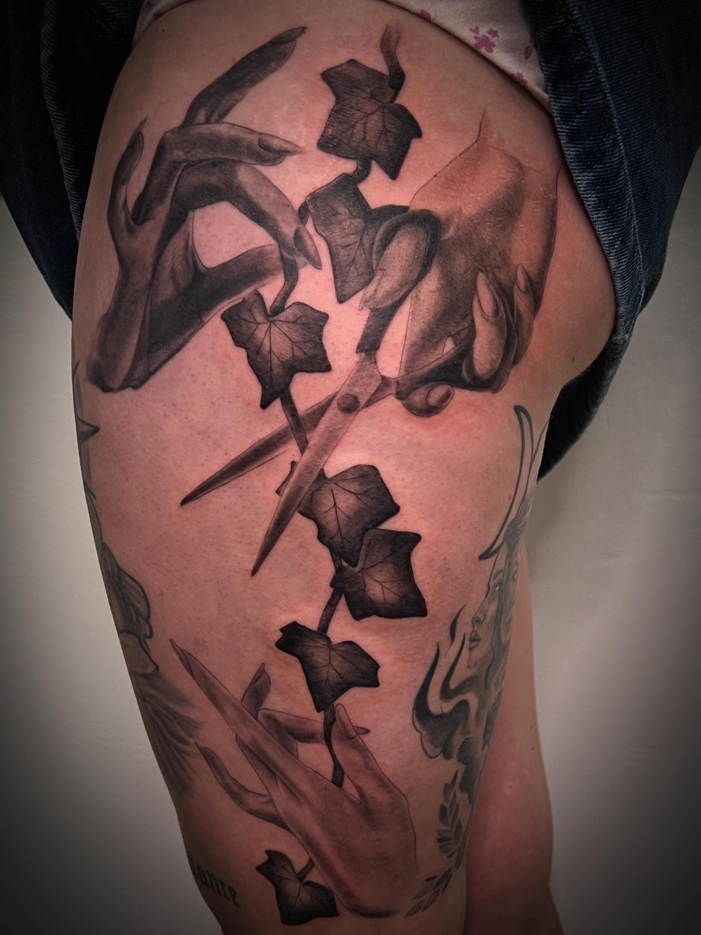 Tattoo of a hand holding a pair of scissors, with leaves and a face depicted in black and gray ink on the thigh.