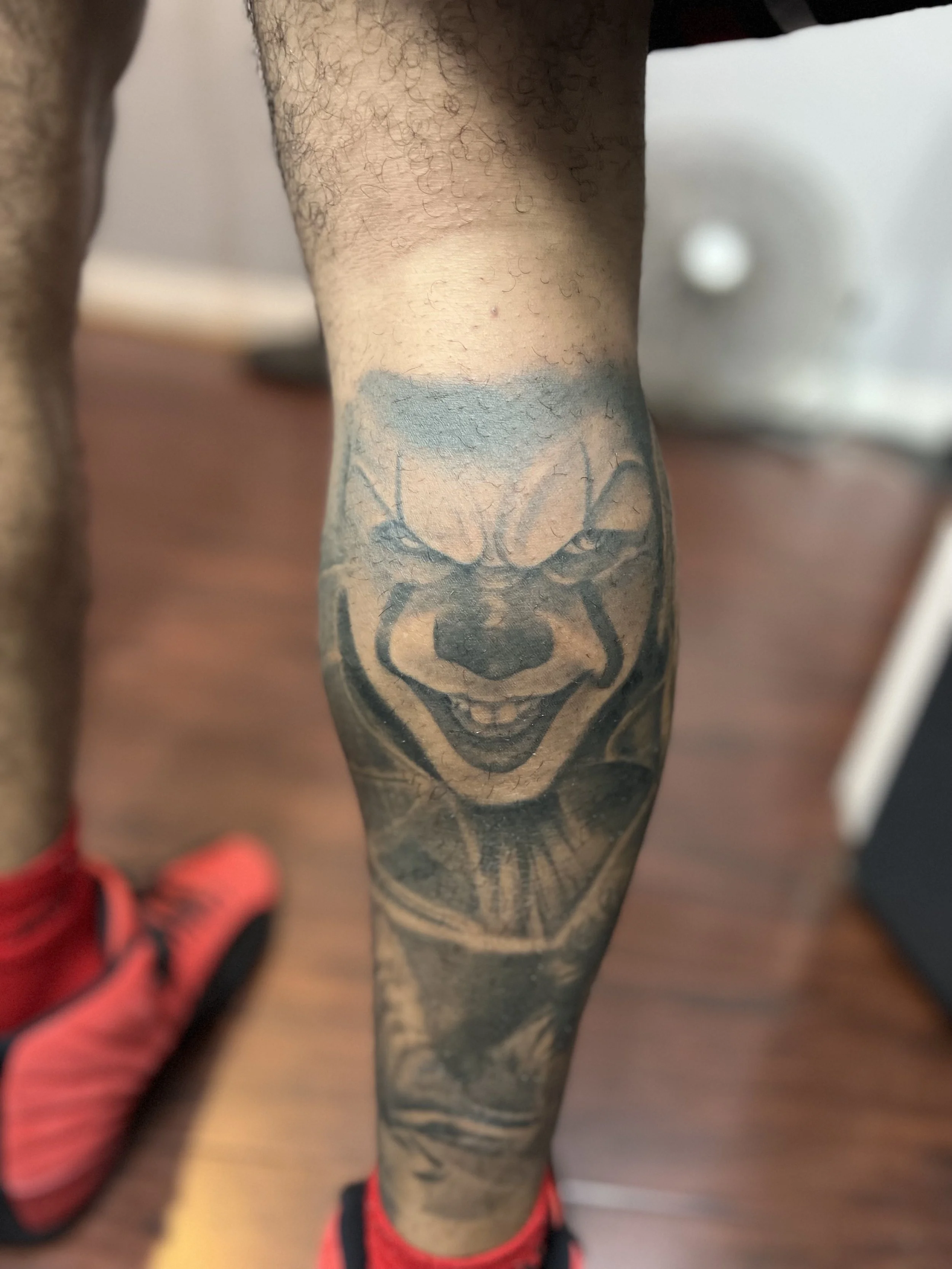 Tattoo on person's lower leg depicting a smiling clown face with open mouth, detailed shading, and a watercolor background.
