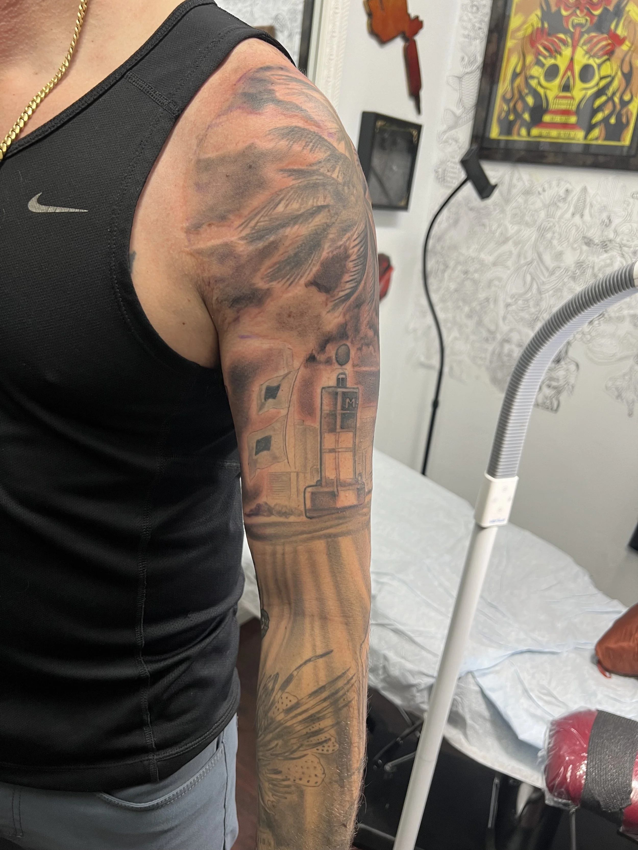 A detailed tattoo sleeve on a person's upper arm, featuring a lighthouse, sailboats, and a sunset sky with clouds and a sun.