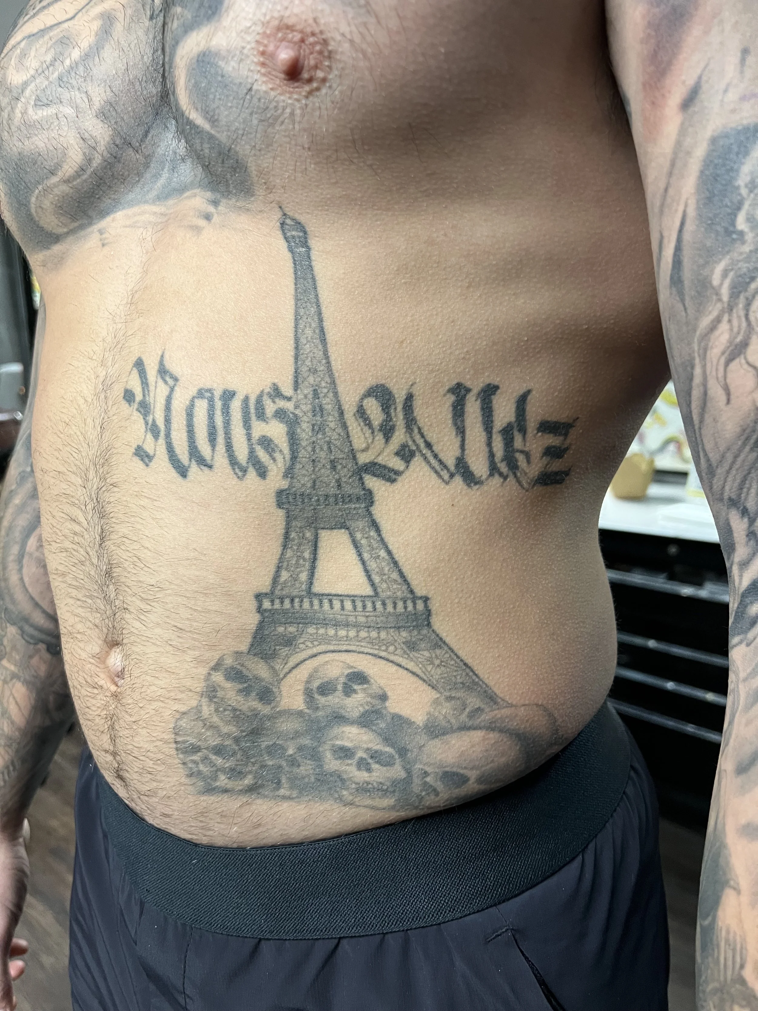 Tattoo of the Eiffel Tower with the words 'Always' above it and skulls at the base on a man's chest and torso.