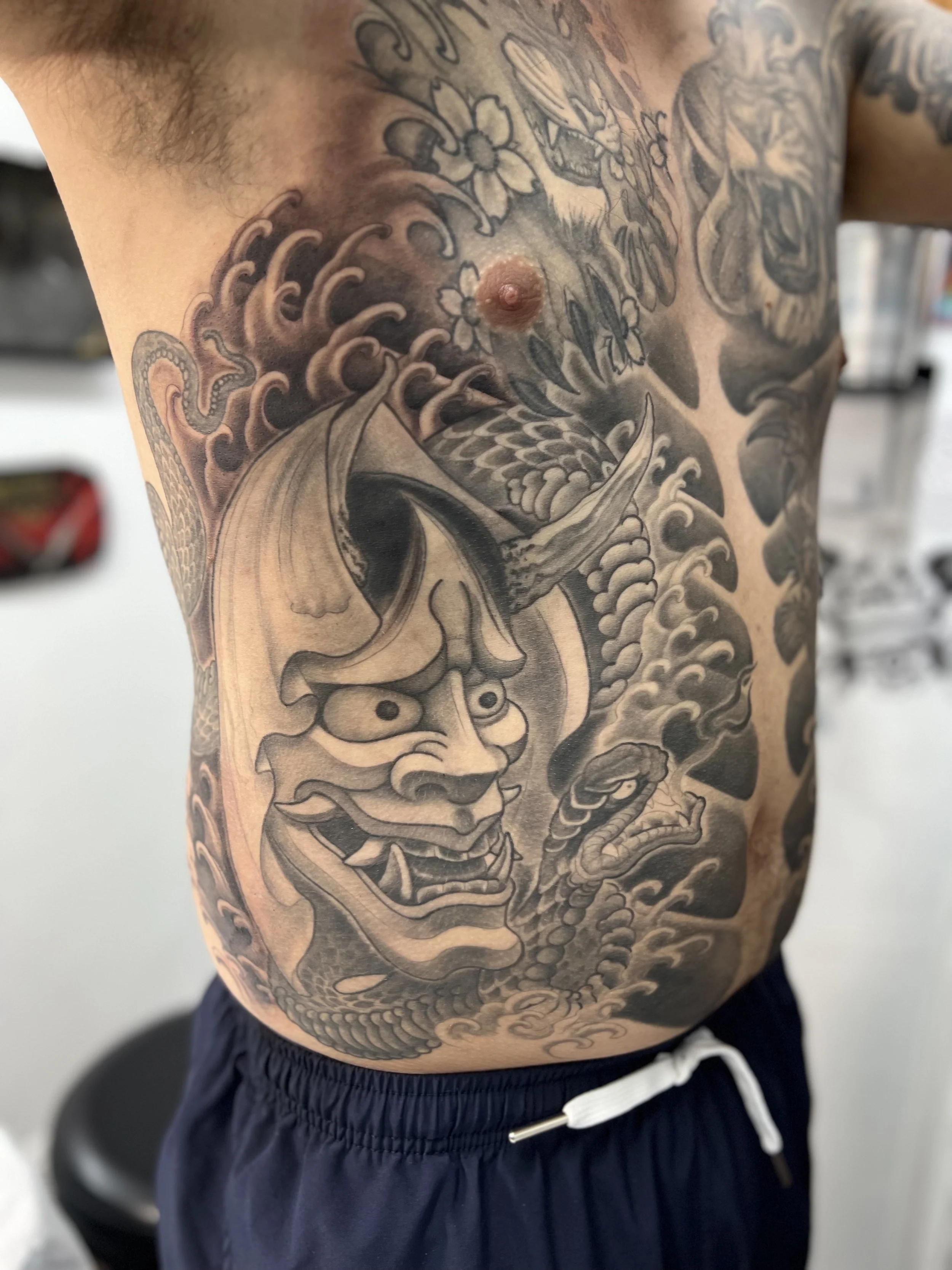 A person's torso with a detailed black and gray Japanese-style tattoo featuring a fierce oni mask, a dragon, and waves.