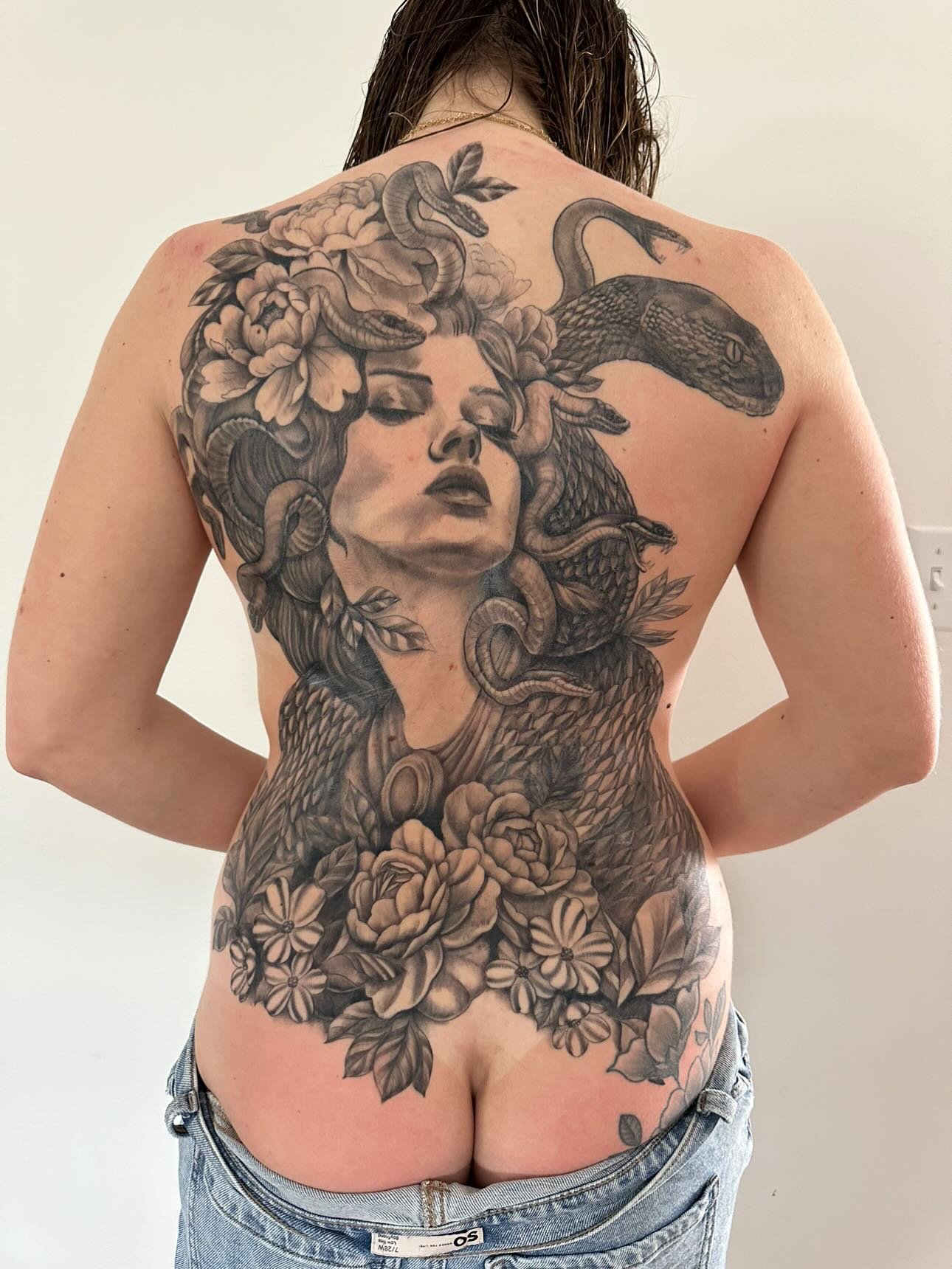 Back view of a person with a large monochrome tattoo of a woman's face, flowers, peacock feathers, and snakes spanning from shoulders to lower back.