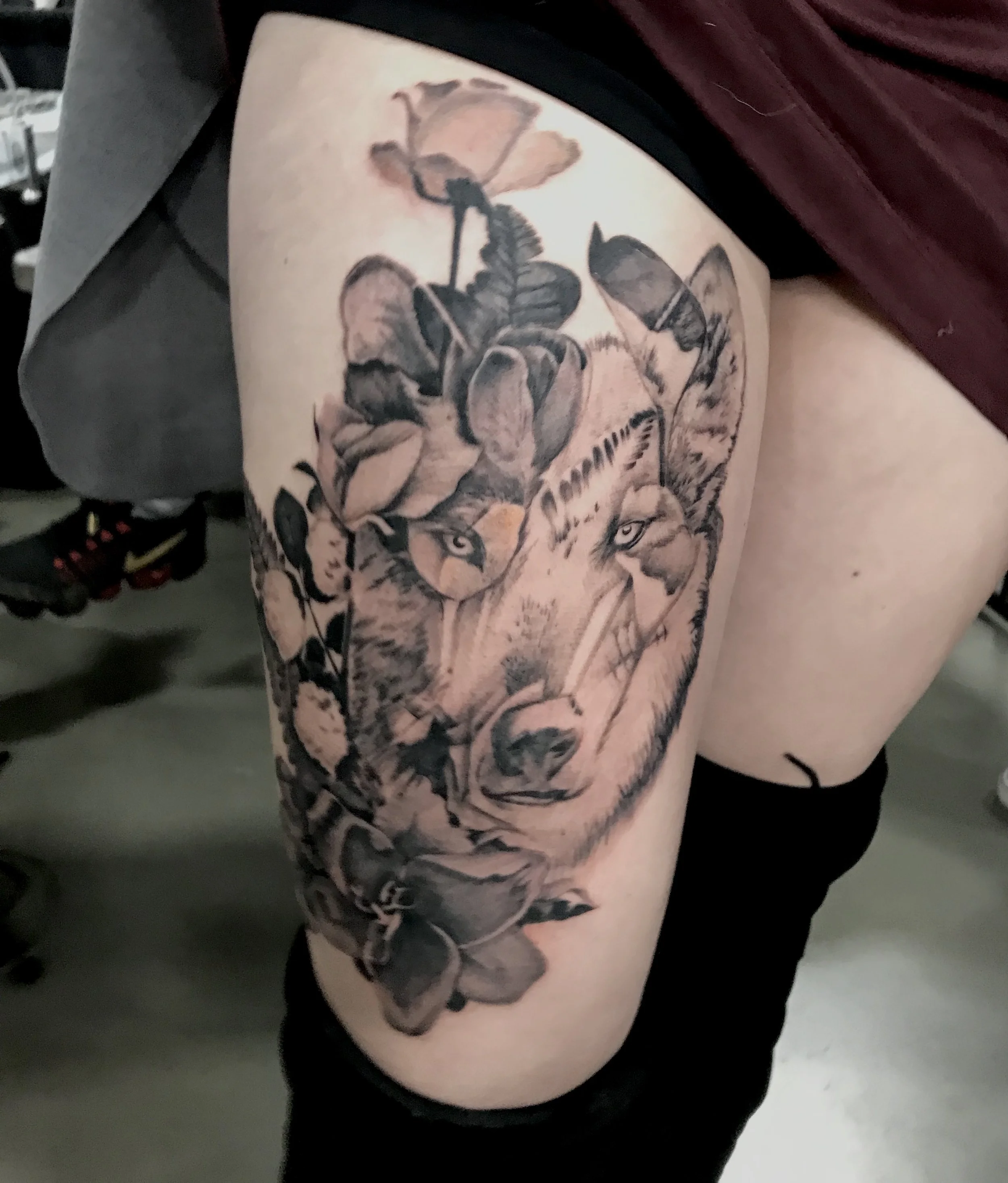 Black and gray tattoo of a wolf's face surrounded by leaves and flowers on a person's thigh.