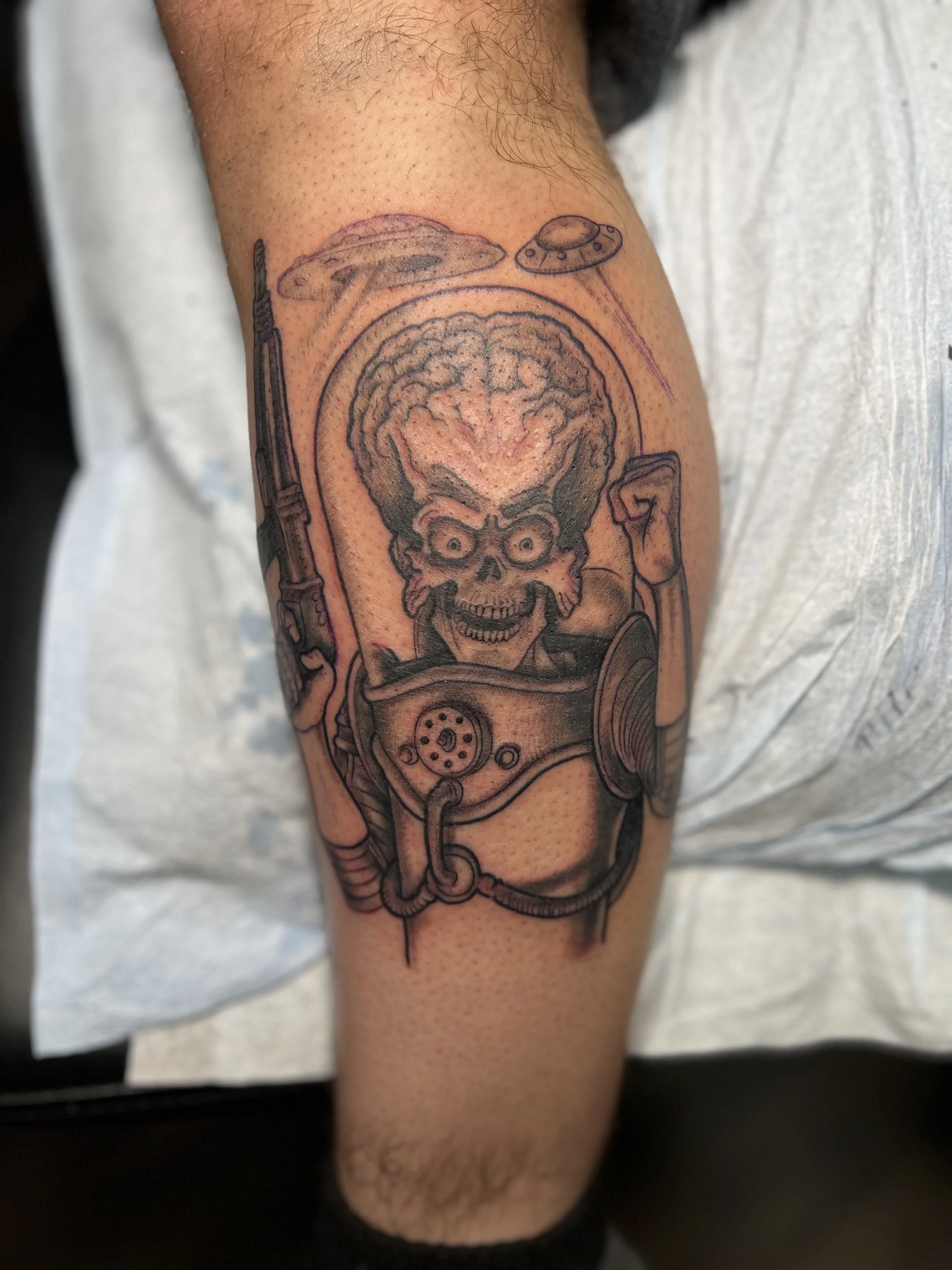 Tattoo of a skeleton with an exposed brain, wearing a space suit, holding a gun, with UFOs in the background.