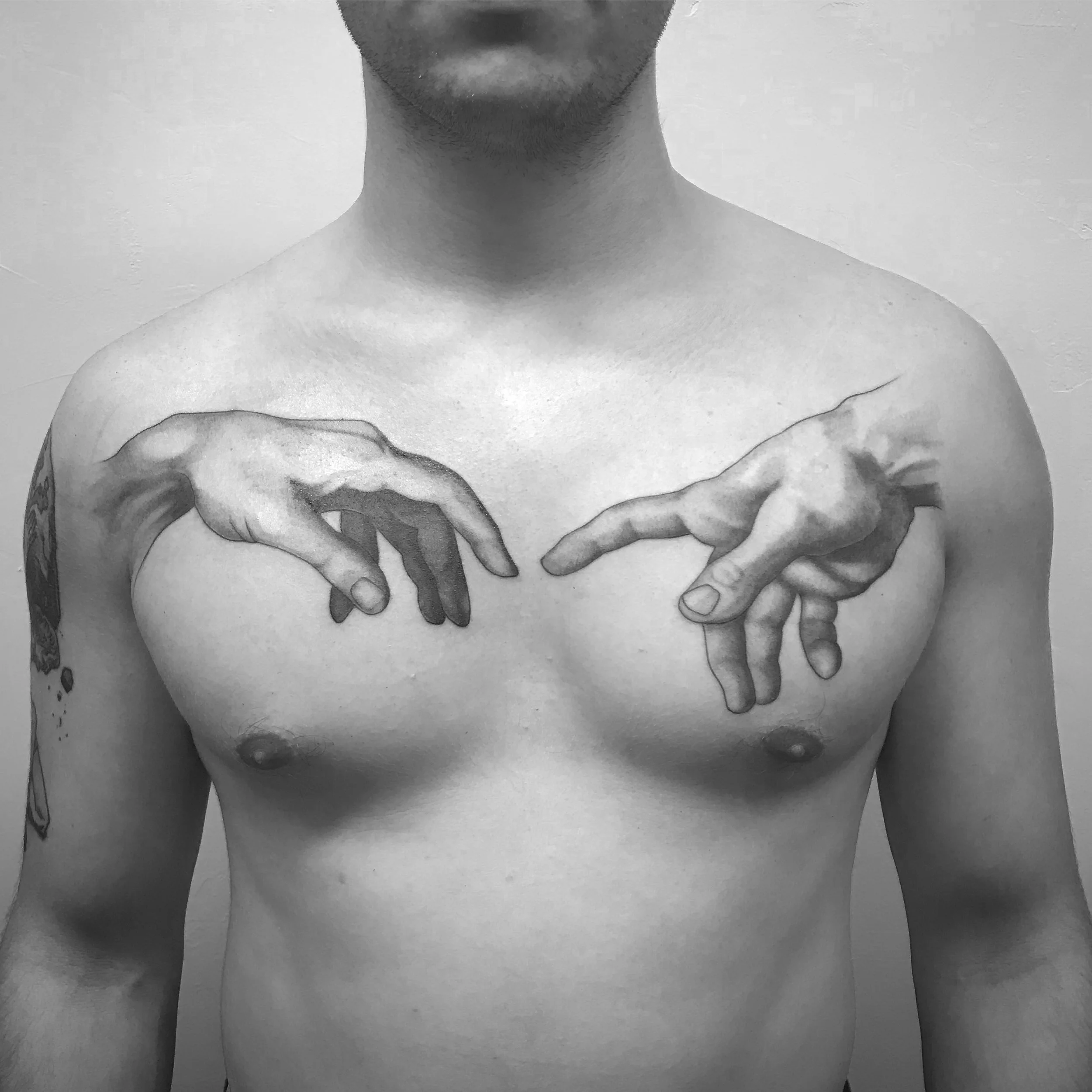 Black and white photo of a man's bare chest with a tattoo of Michelangelo's 'The Creation of Adam' on his upper chest.