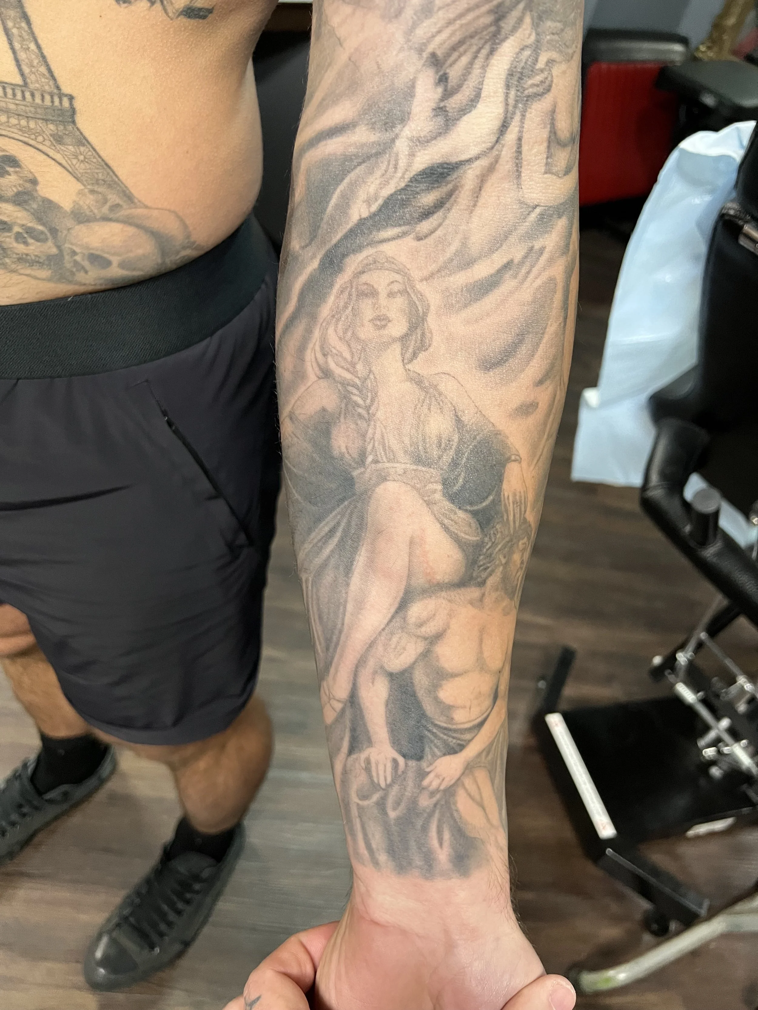 A black and gray tattoo on a person's arm depicting a woman with long hair and a braid, sitting on a muscular man's shoulders, with swirling clouds or smoke around her.