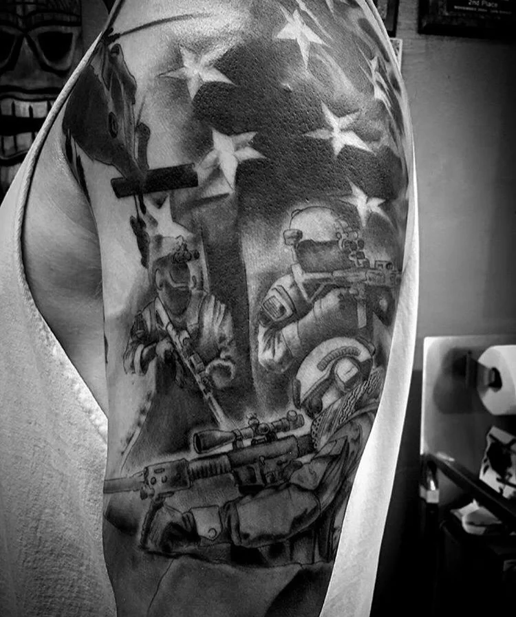 Black and white tattoo on upper arm featuring soldiers, military gear, and the American flag in the background.