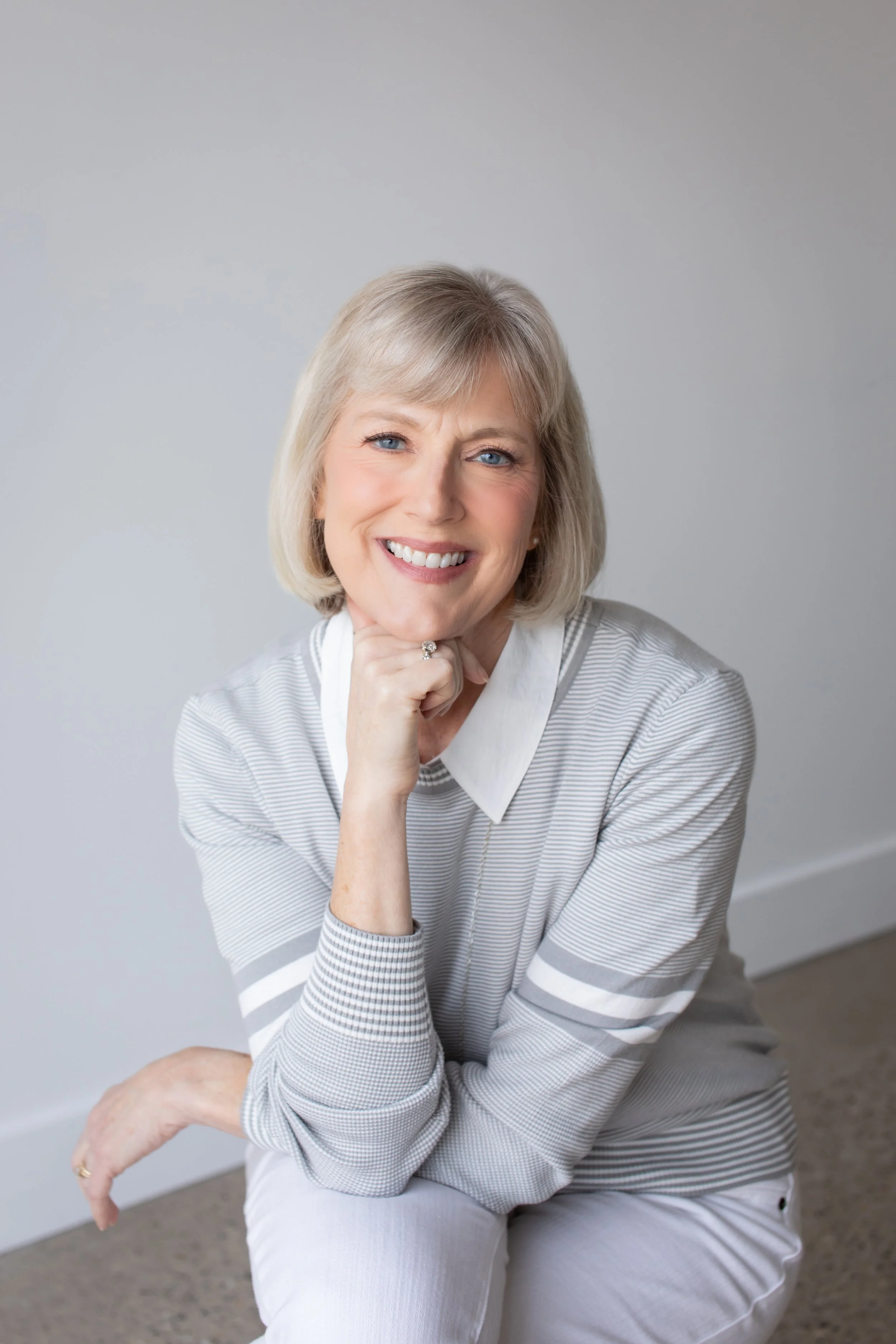 A smiling woman with short blonde hair, wearing a gray and white striped sweater, sitting on a chair in a room with a plain gray wall.
