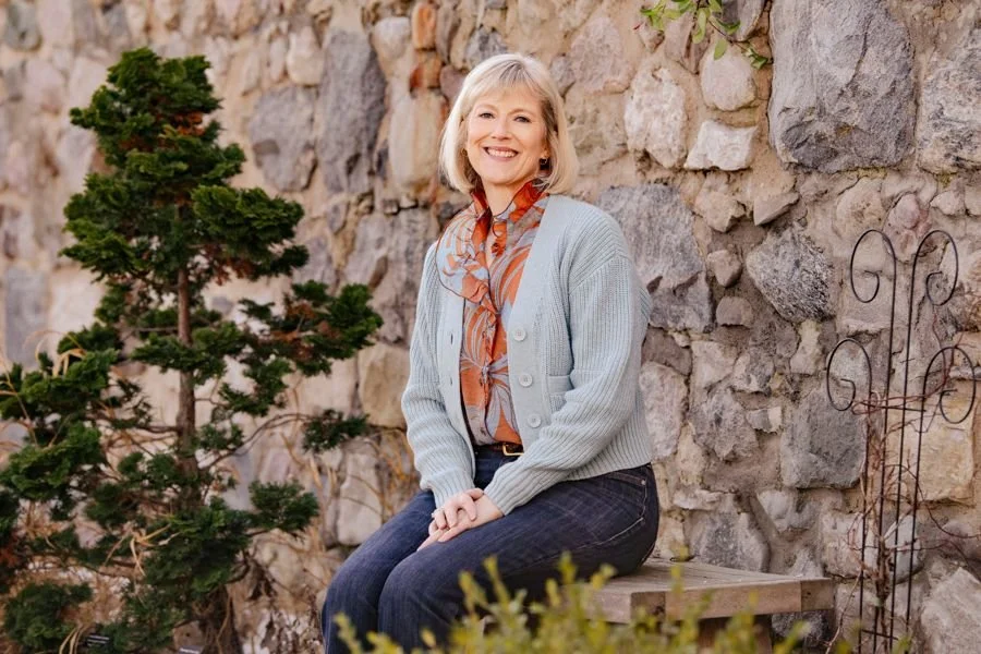 Mary Pate, founder of Mustard Seed Health Coaching and the SheSTANDS Method, smiling in a light blue cardigan, seated on a wooden bench beside a stone wall.