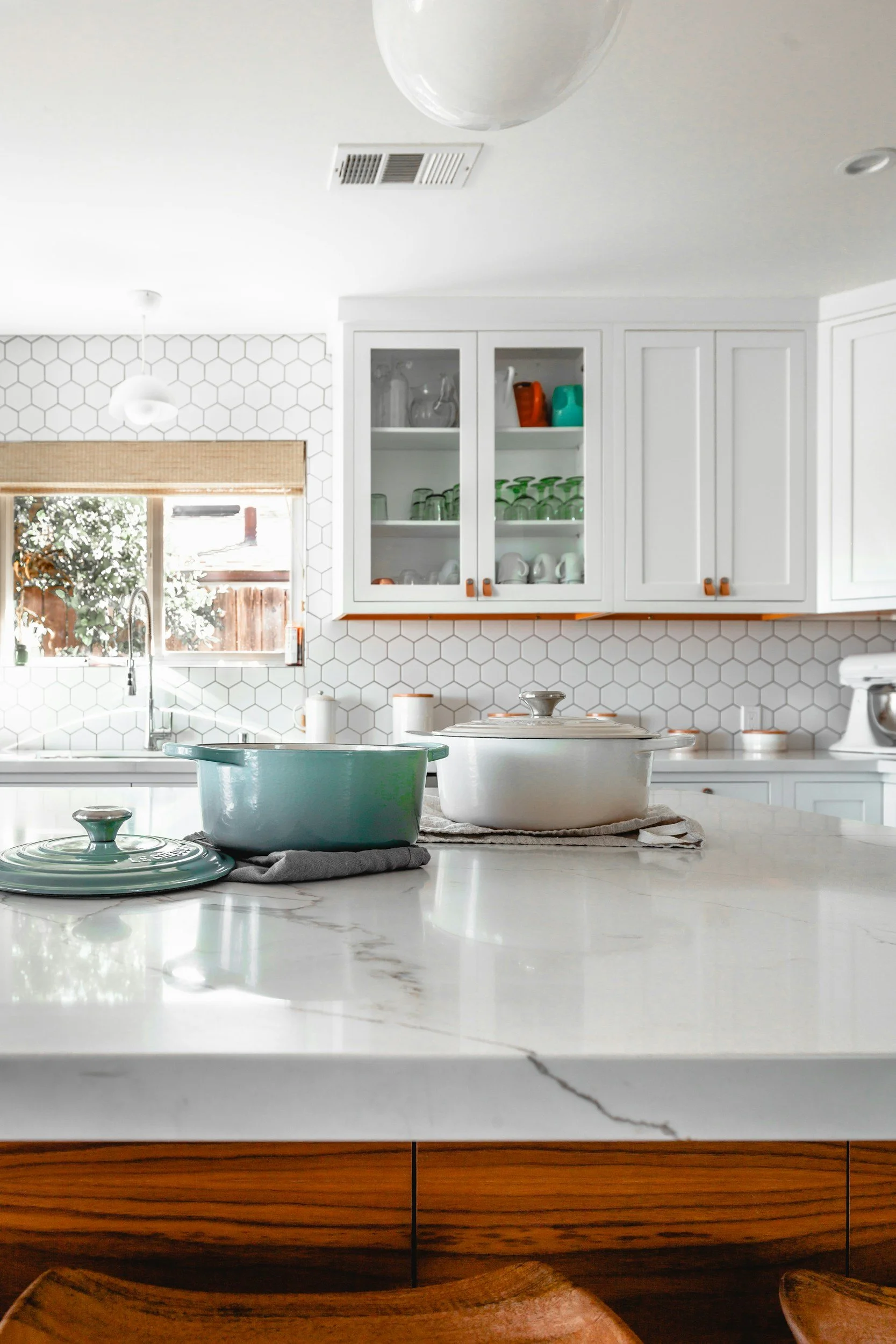 Kitchen with white hexagon tiled backsplash, white upper cabinets with glass doors, and a marble countertop with cooking pots, a mug, and a bowl. There is a window with a bamboo shade, and a wooden dining table with chairs in the foreground.