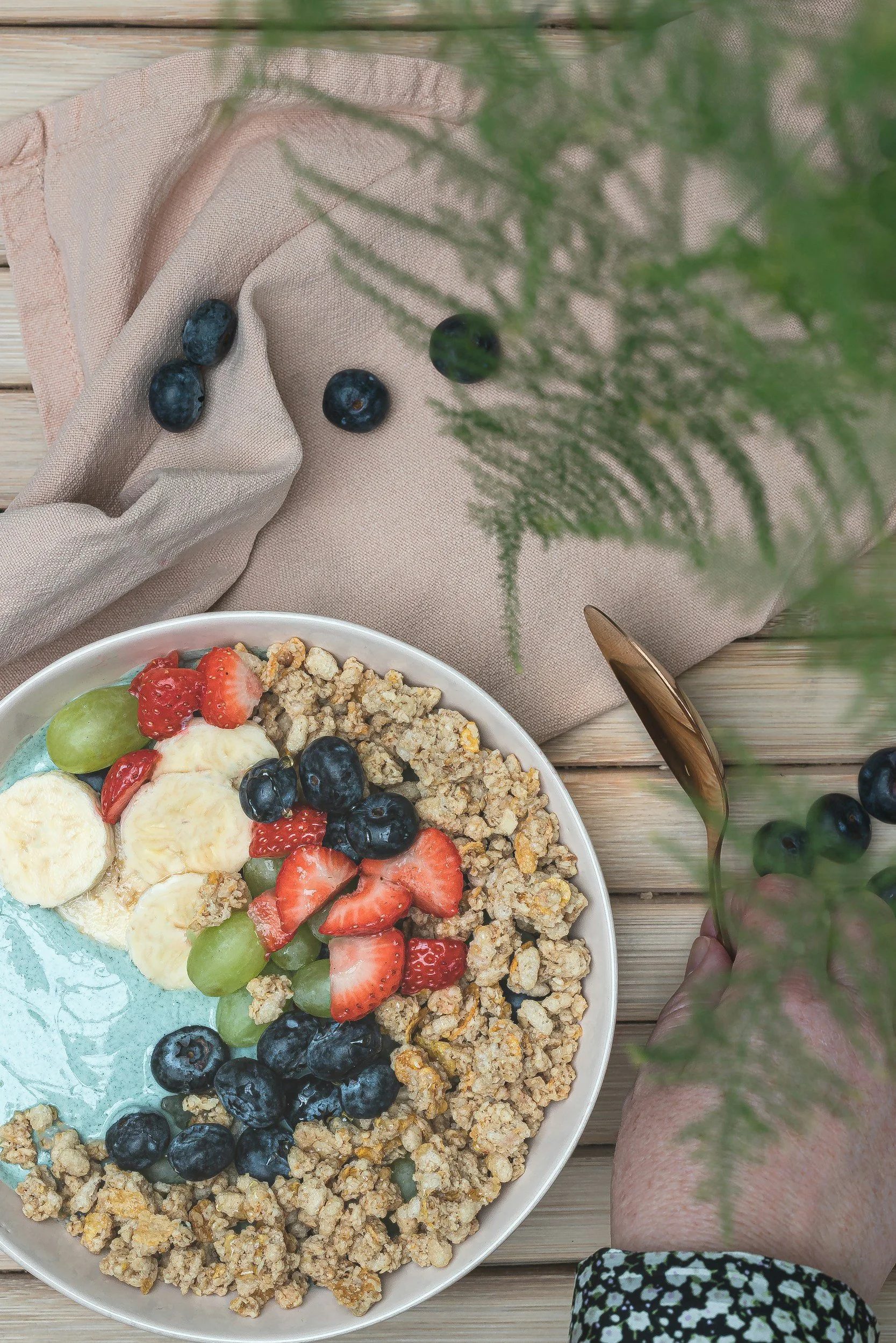 A bowl of granola topped with sliced bananas, strawberries, blueberries, green grapes, and yogurt, placed on a wooden table with a beige cloth and a green plant nearby.