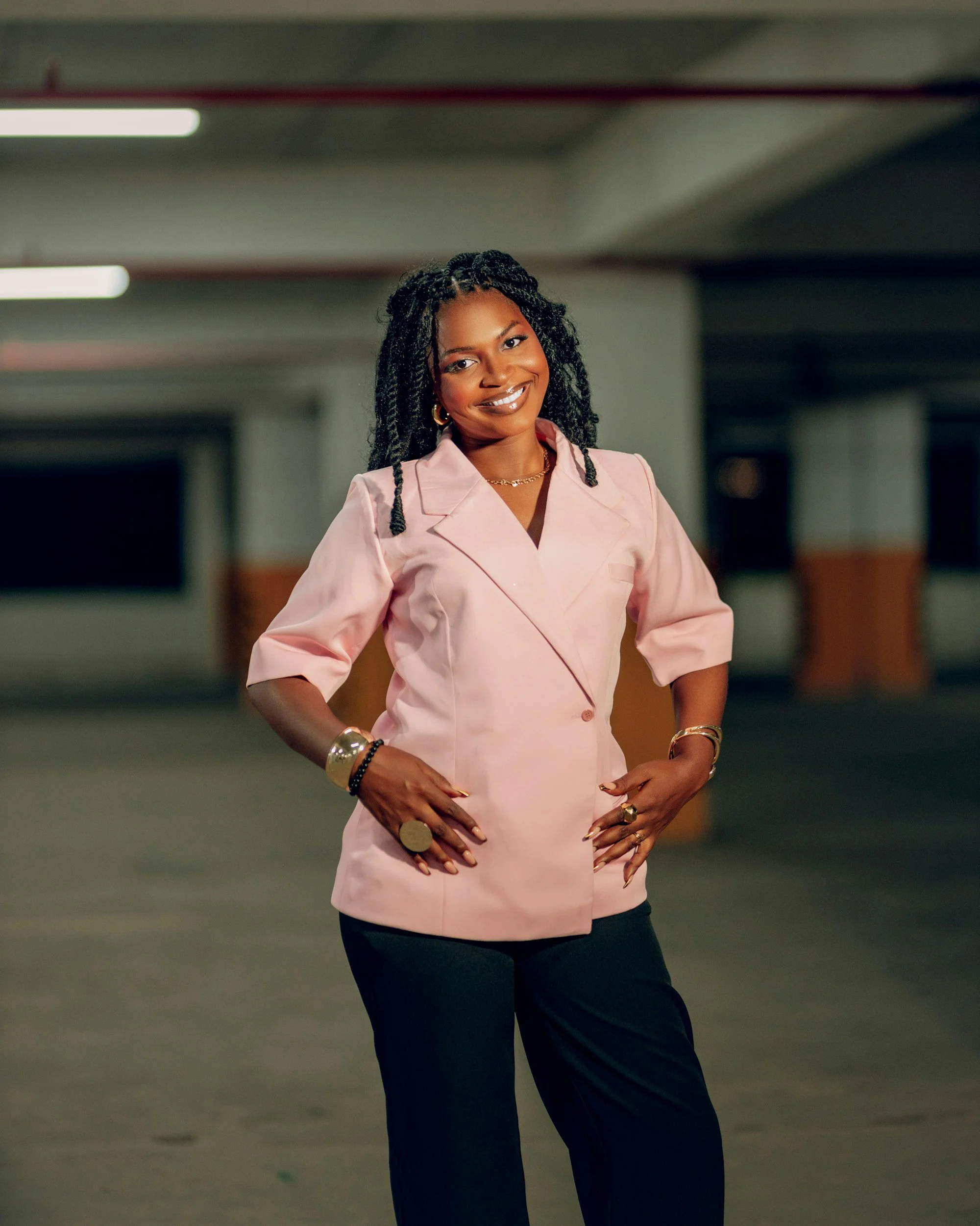 Woman with dark skin and curly black hair styled in twists, smiling confidently, standing in a dimly lit parking garage, wearing a light pink blazer, black pants, and accessorized with gold jewelry.