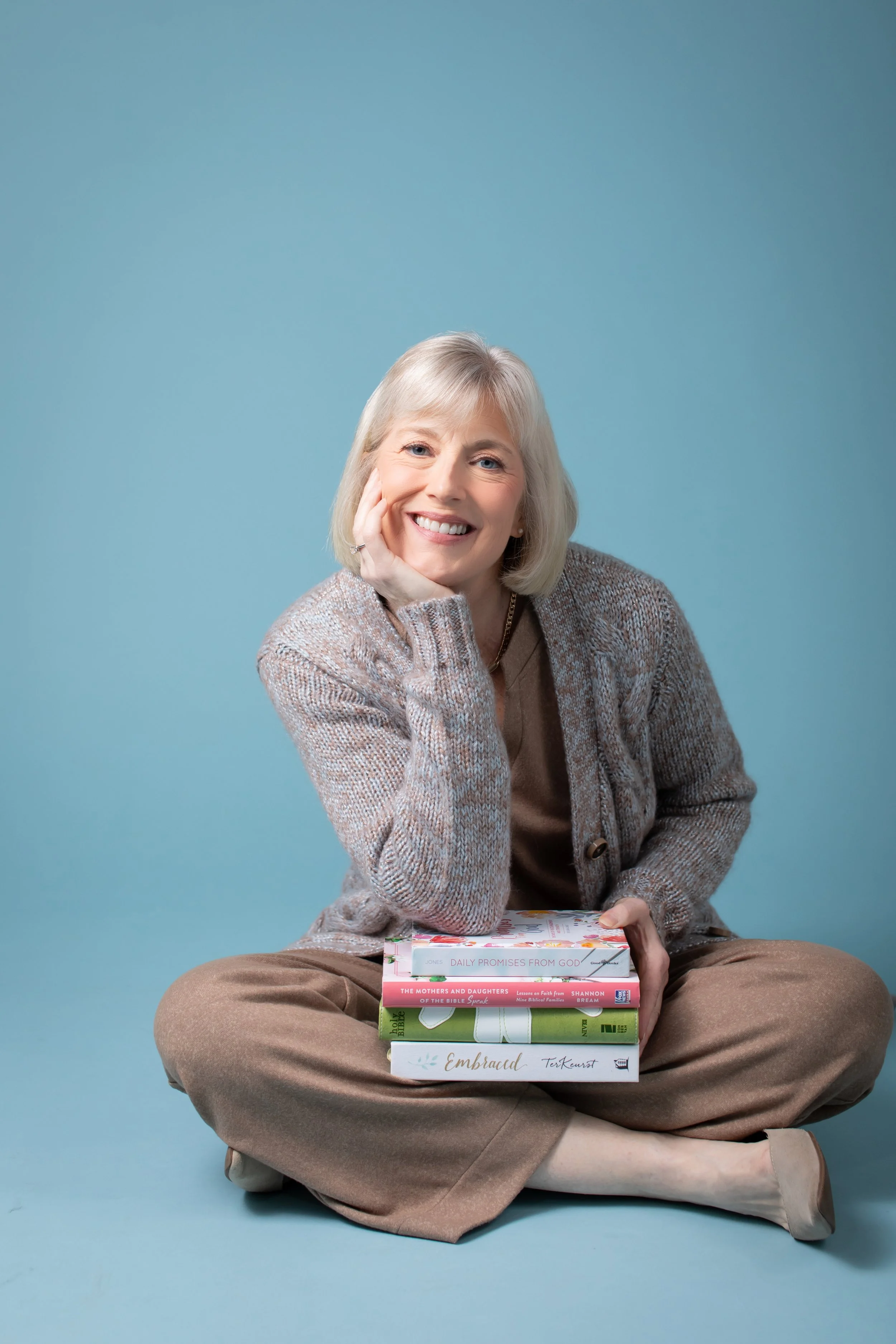 A smiling elderly woman with gray hair, wearing a brown shirt, beige cardigan, and brown pants, sitting cross-legged against a blue background. She rests her head on her left hand and has a stack of five books on her lap, titled from top to bottom: "Daily Promises from God," "The Mothers and Daughters of the Bible Speak," "Lessons on Faith from Nine Biblical Families," "The Bible and the Holy Spirit," and "Embraced."