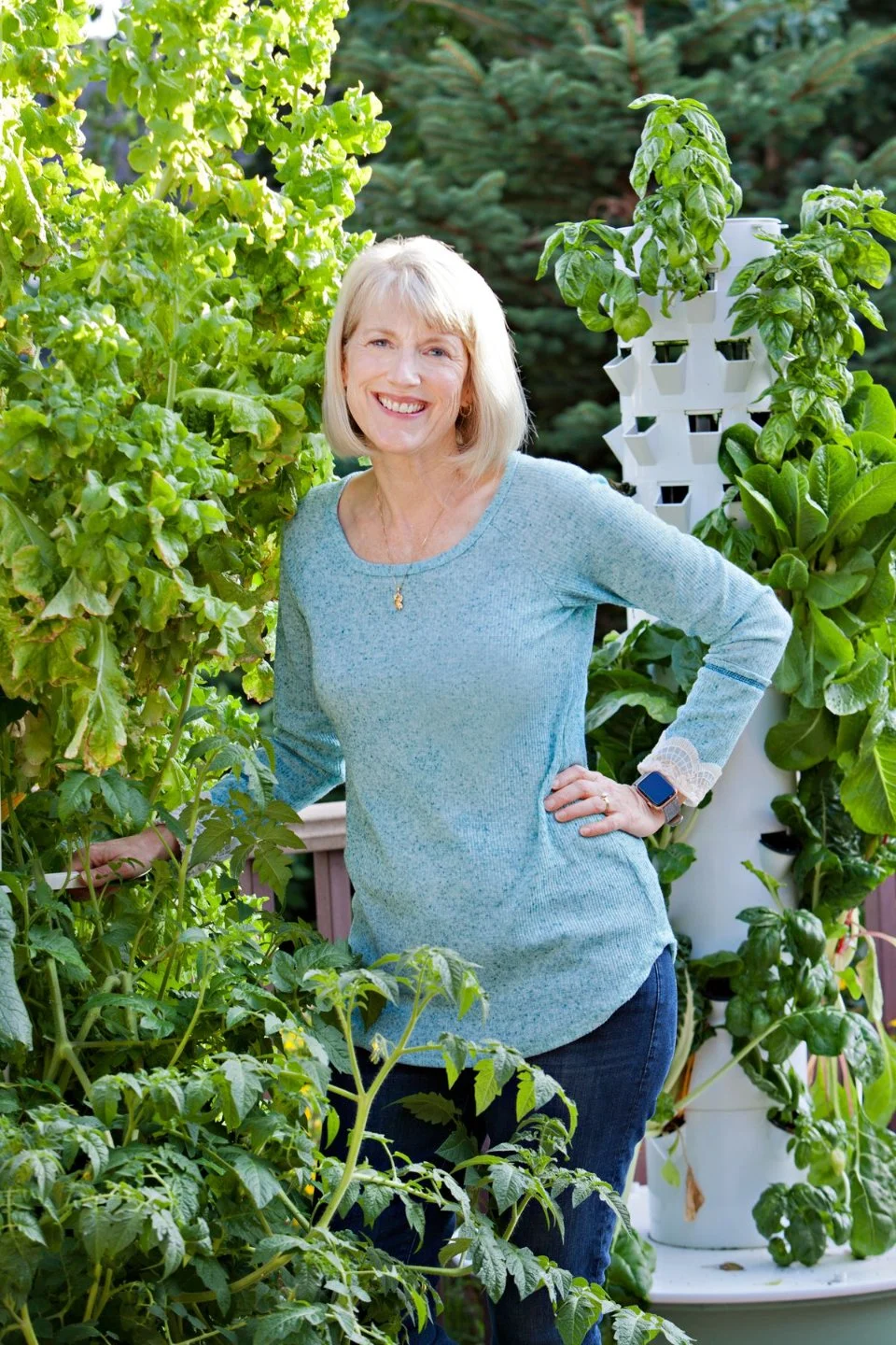 A smiling woman with blonde hair wearing a blue long-sleeve top and jeans, standing in a lush garden with various green plants, including tomato and basil, and a vertical garden planter in the background.