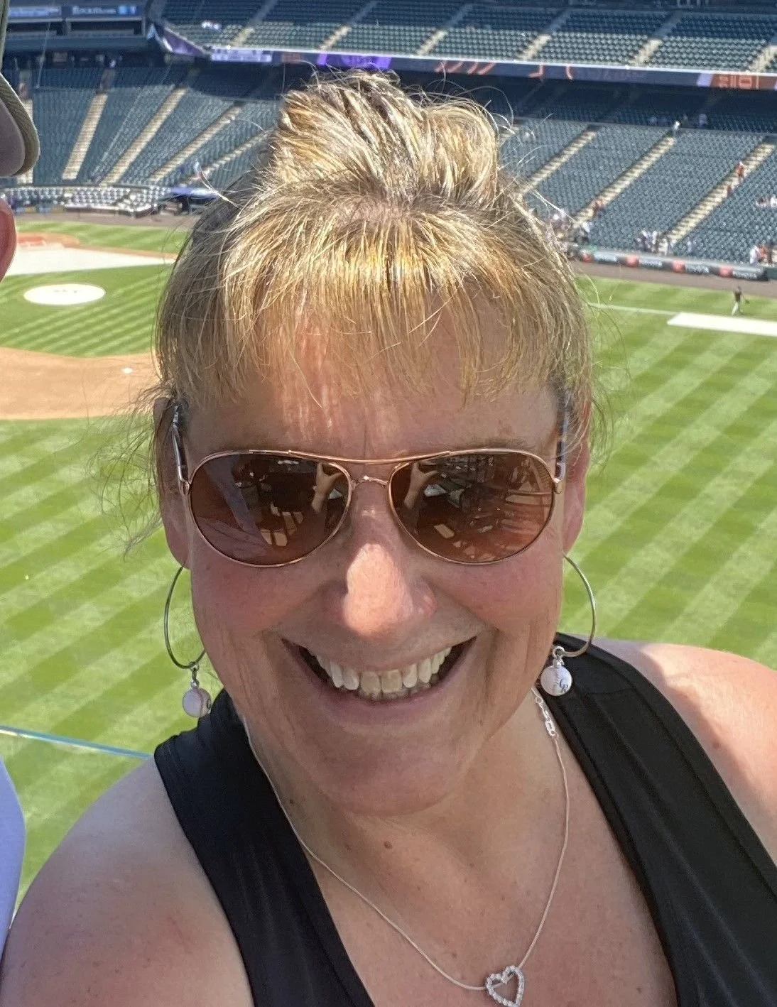 A woman with blonde hair in a bun, wearing sunglasses, earrings, and a black top, smiling for a selfie at a baseball stadium.