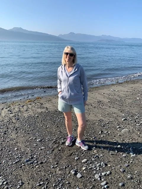 Mary Pate, founder of Mustard Seed Health Coaching, standing on a rocky beach in Homer, Alaska, with mountains and water behind her.