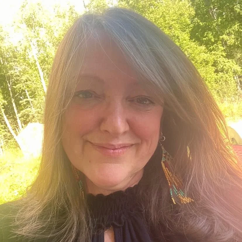 A woman with long, light brown hair standing outdoors with green trees in the background, smiling at the camera during daytime.