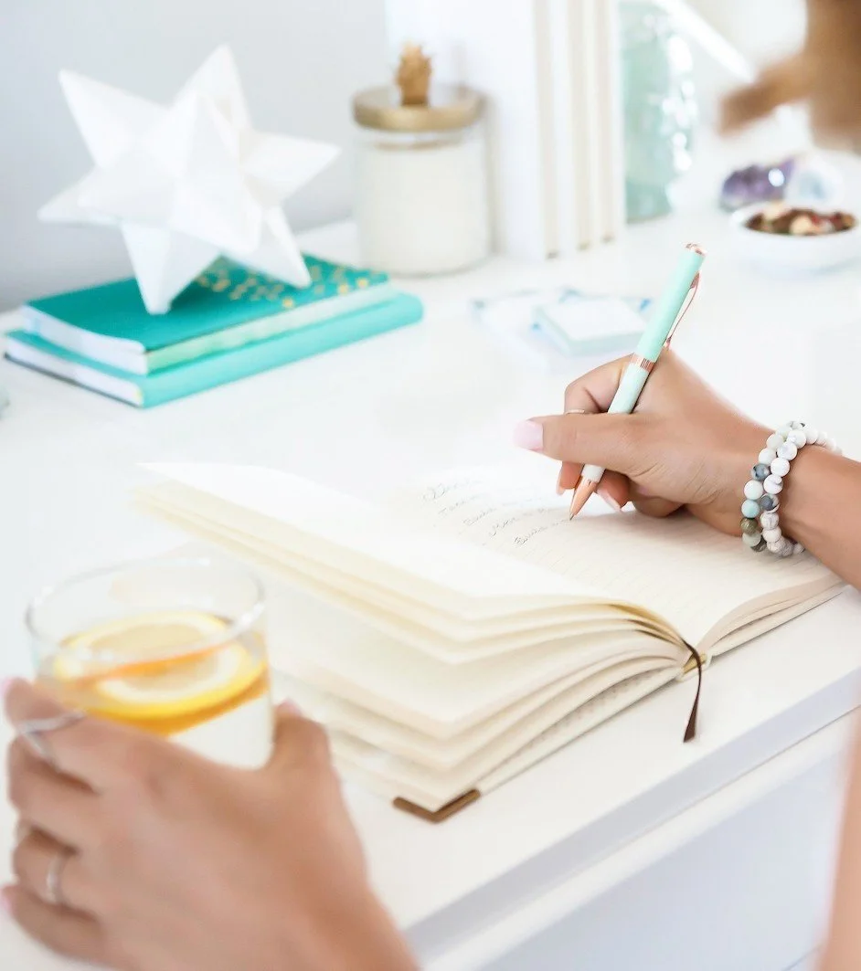 Person writing in notebook with a pen, holding a glass of lemon water, on a white desk with decorative items including a star-shaped paper lantern and teal books.