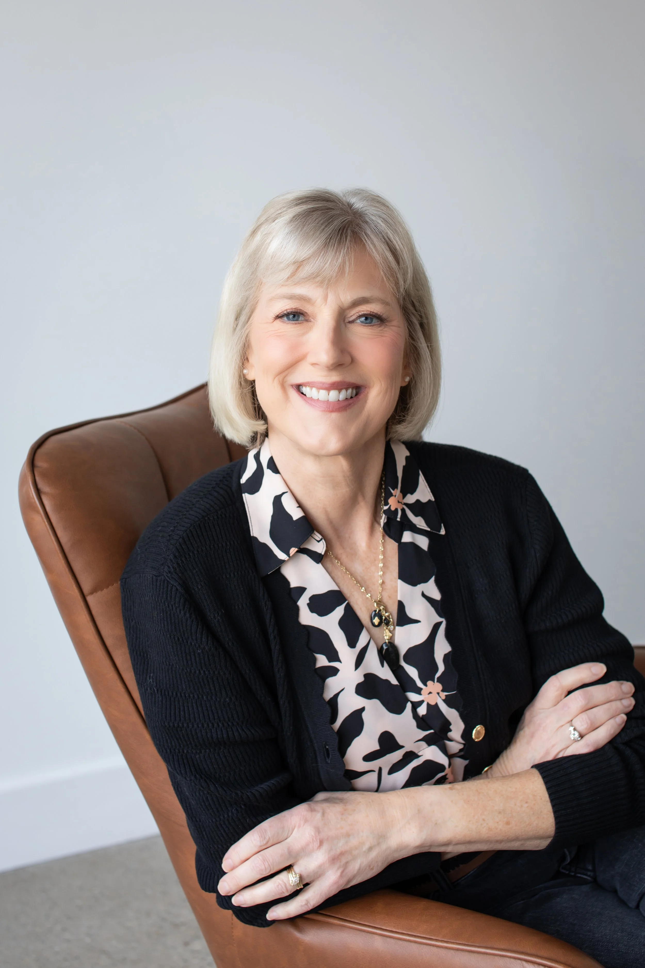 Mary Pate, founder of Mustard Seed Health Coaching and the SheSTANDS Method, smiling in a black cardigan and floral blouse, seated in a brown leather chair.
