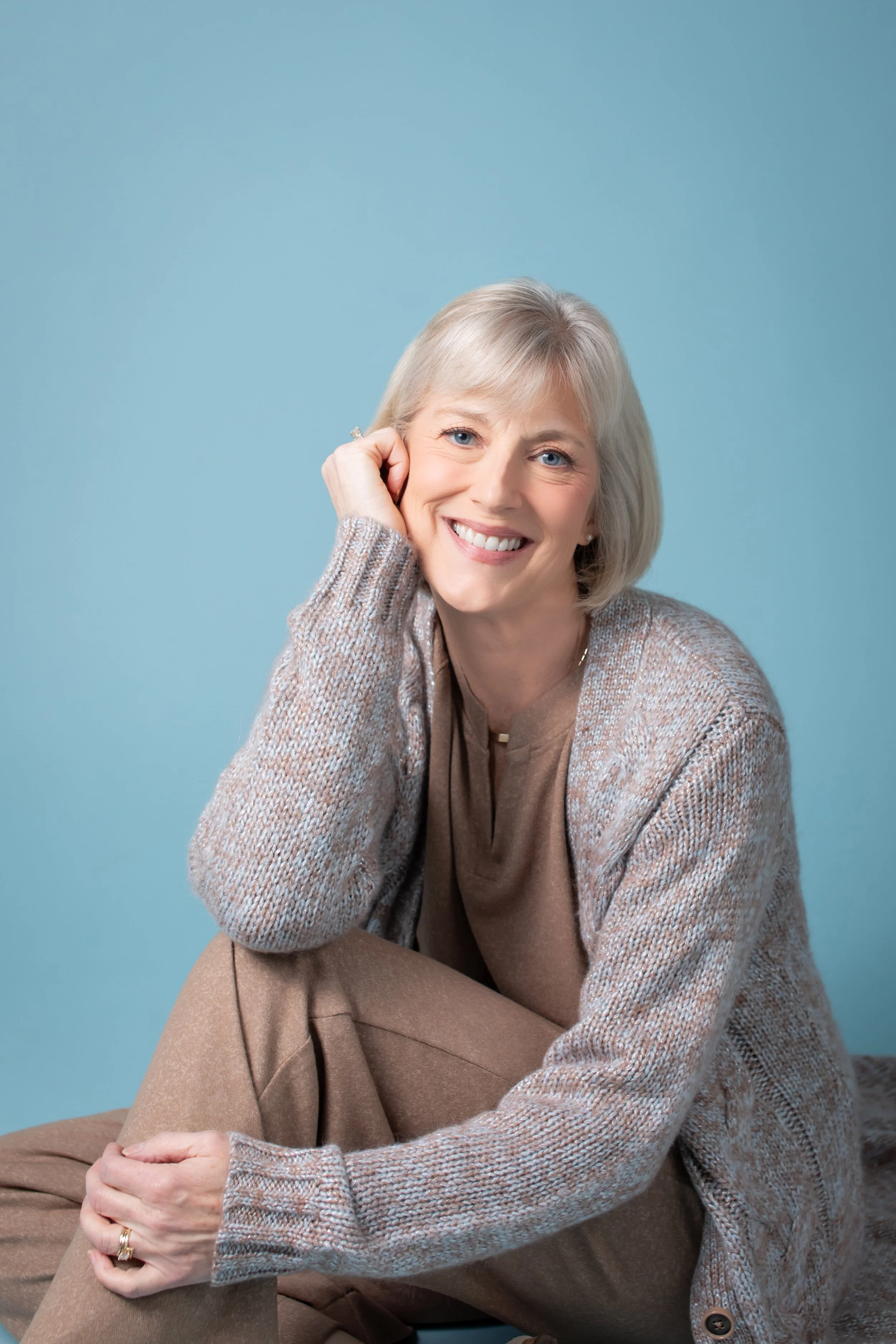 A smiling middle-aged woman with grey hair, blue eyes, and light skin, sitting against a light blue background. She is resting her head on her right hand and wearing a cozy knit sweater in neutral tones.