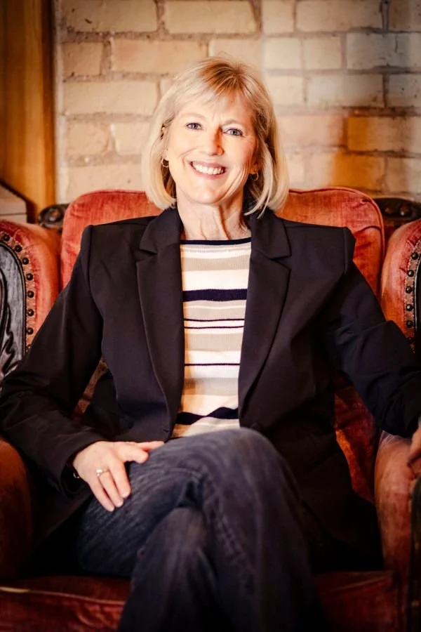 Mary Pate, founder of Mustard Seed Health Coaching and the SheSTANDS Method,  smiling in a navy blazer, seated in a red upholstered chair against a brick wall.