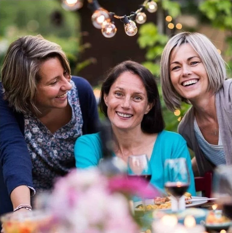 Three women smiling and enjoying outdoor dinner with wine glasses, string lights, and pink flowers.