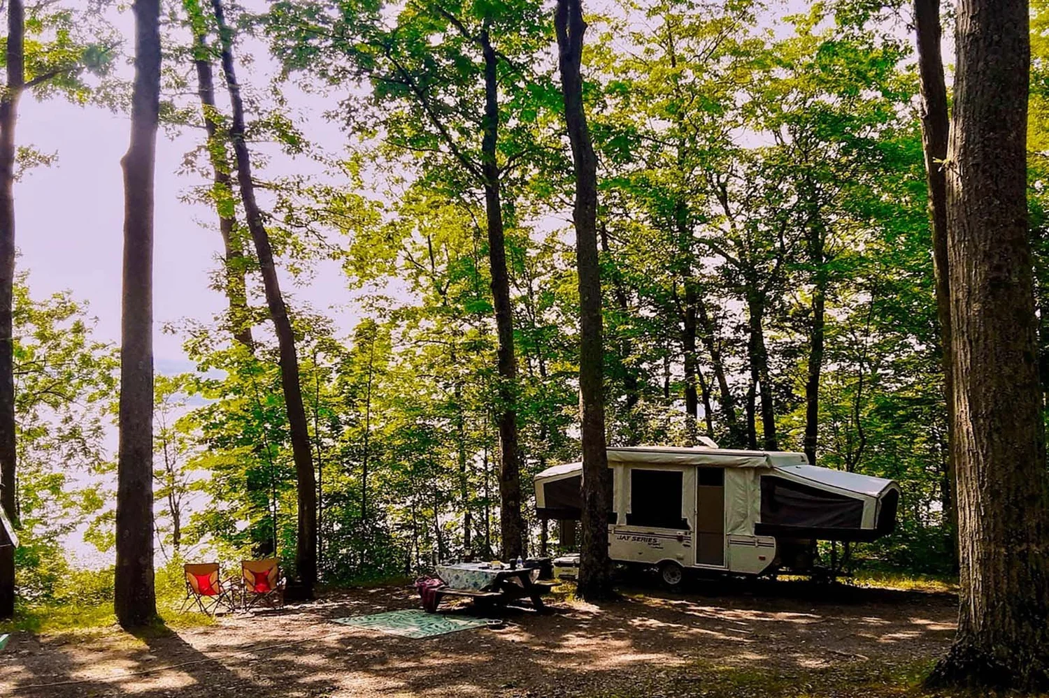 A wooded campsite at Smith Memorial Park with a travel trailer, picnic table, and camp chairs