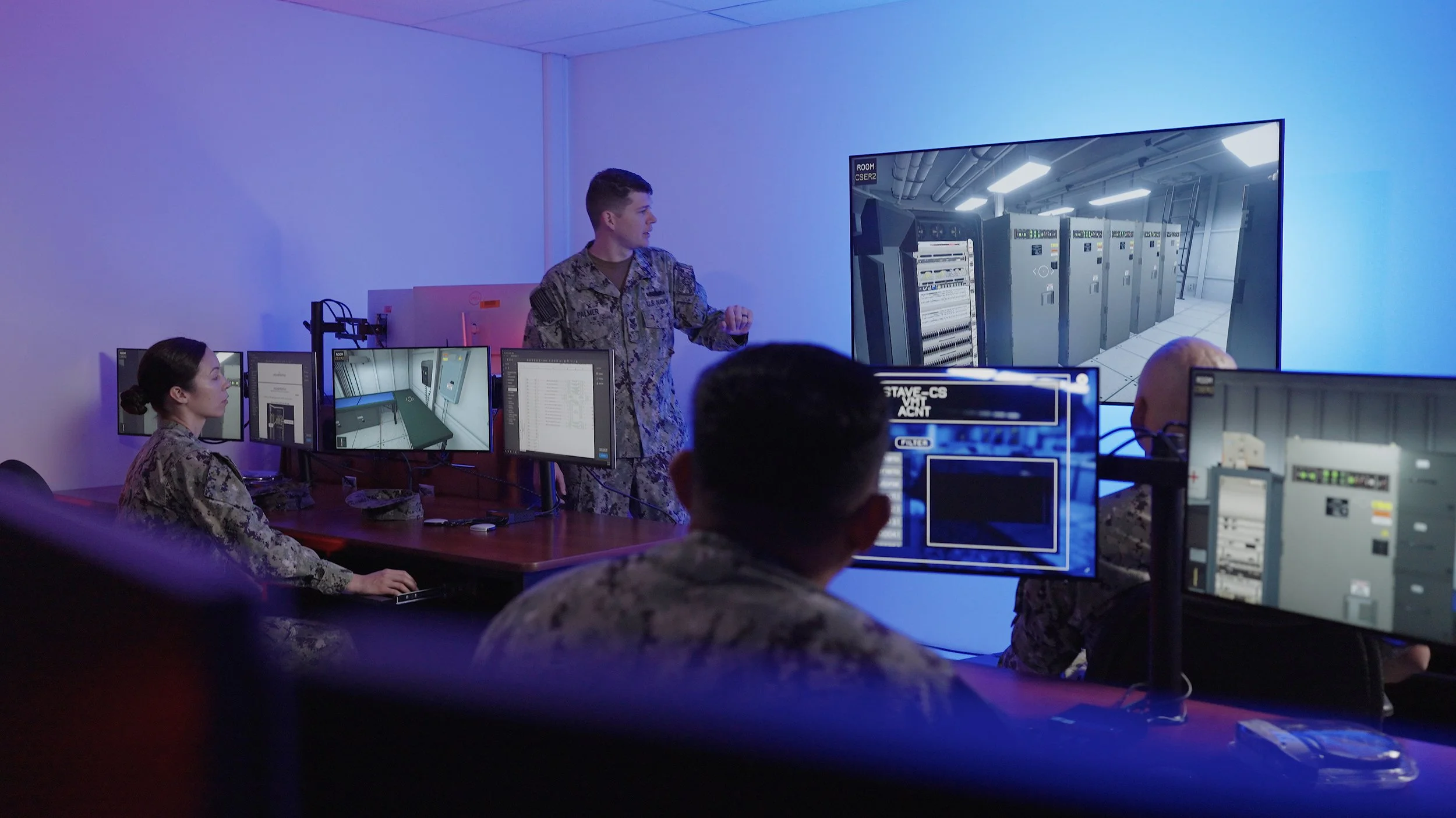 A military personnel in camouflage uniform giving a presentation to a group in a computer room with multiple monitors and a large screen showing a data center or server room.