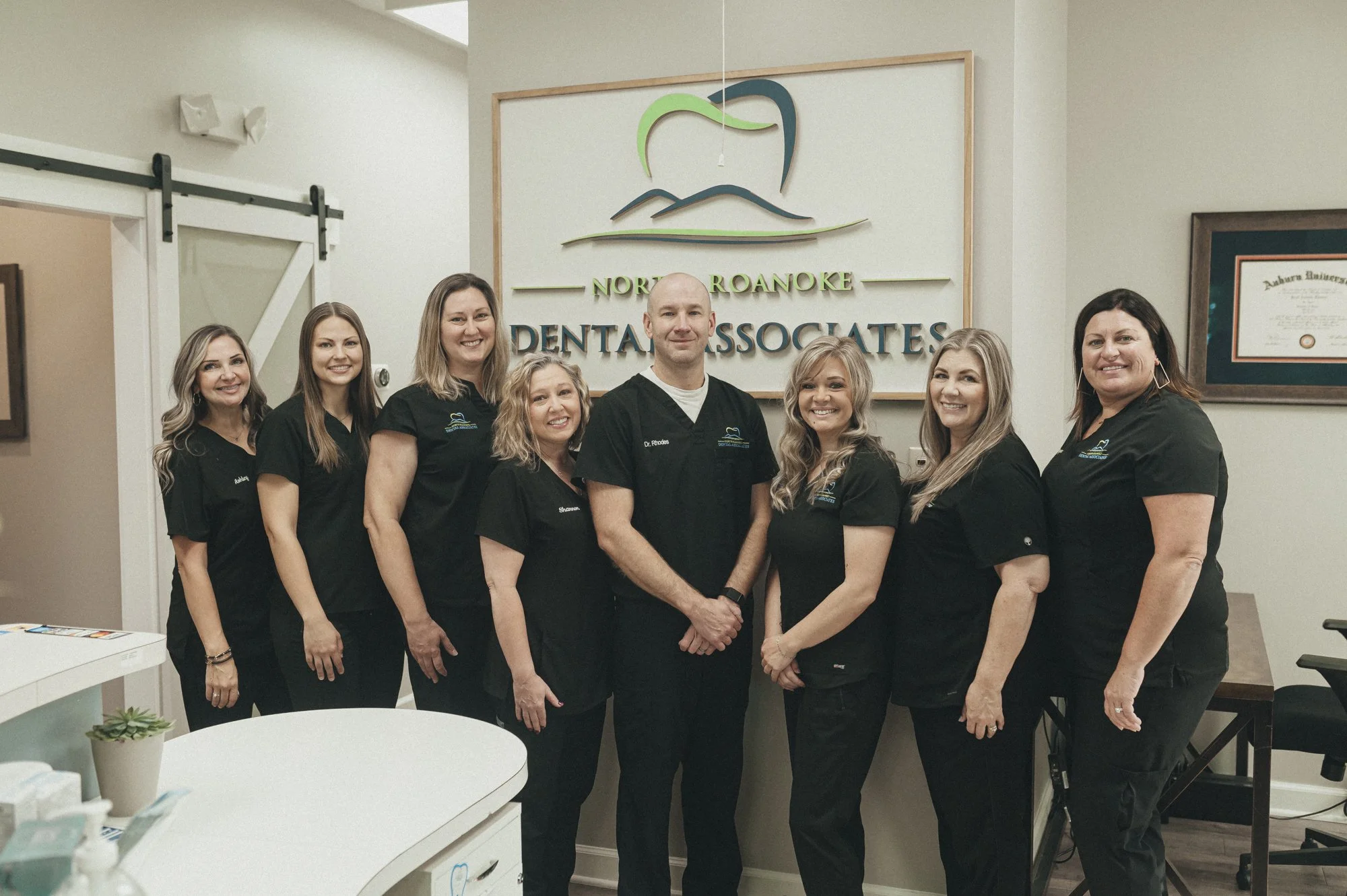 Group of nine dental professionals standing inside a dental office, smiling, wearing black scrubs, in front of a sign for North Roanoke Dental Associates.