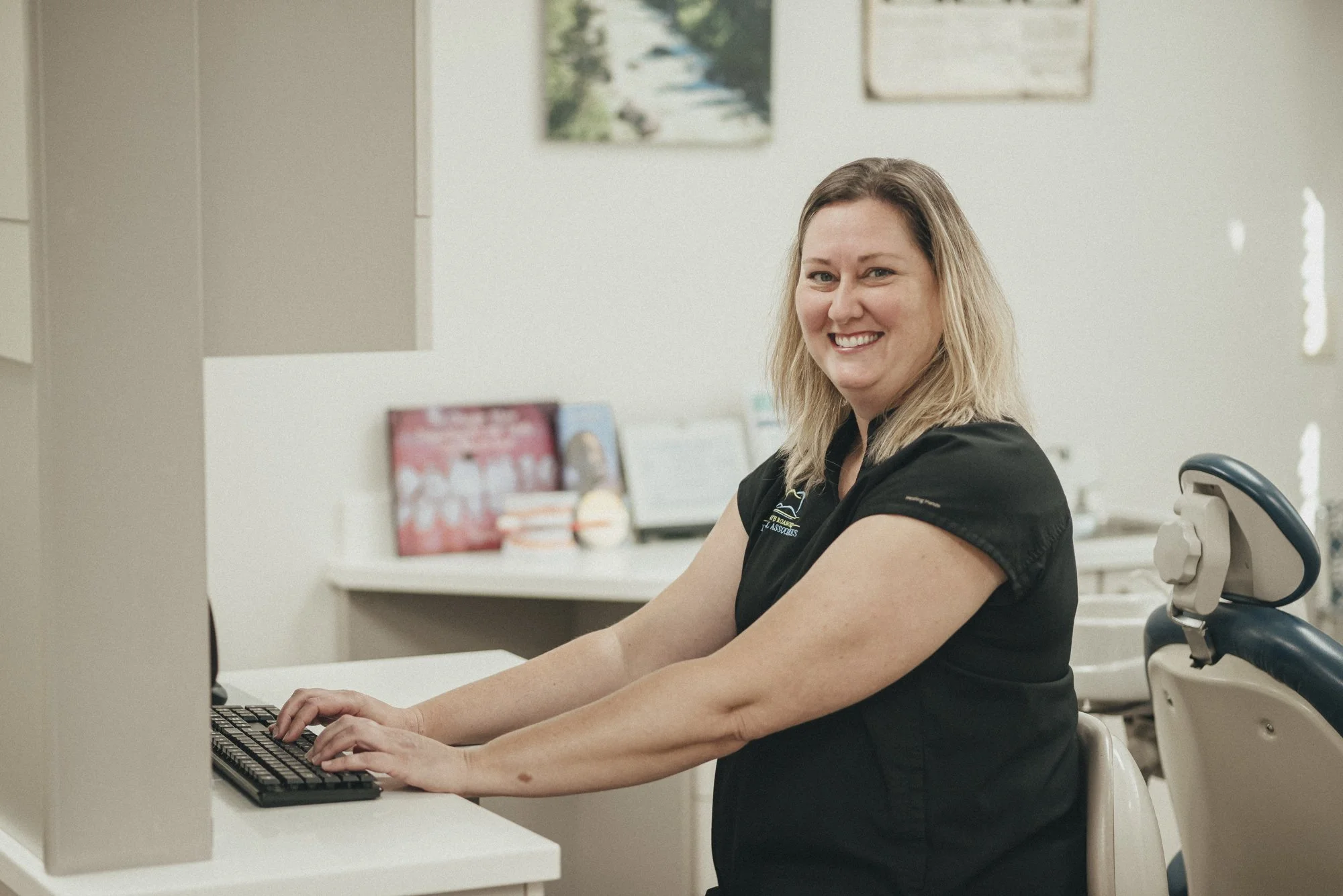 A woman in a medical uniform sitting at a desk, smiling, with a computer keyboard in front of her in a clinical office setting.