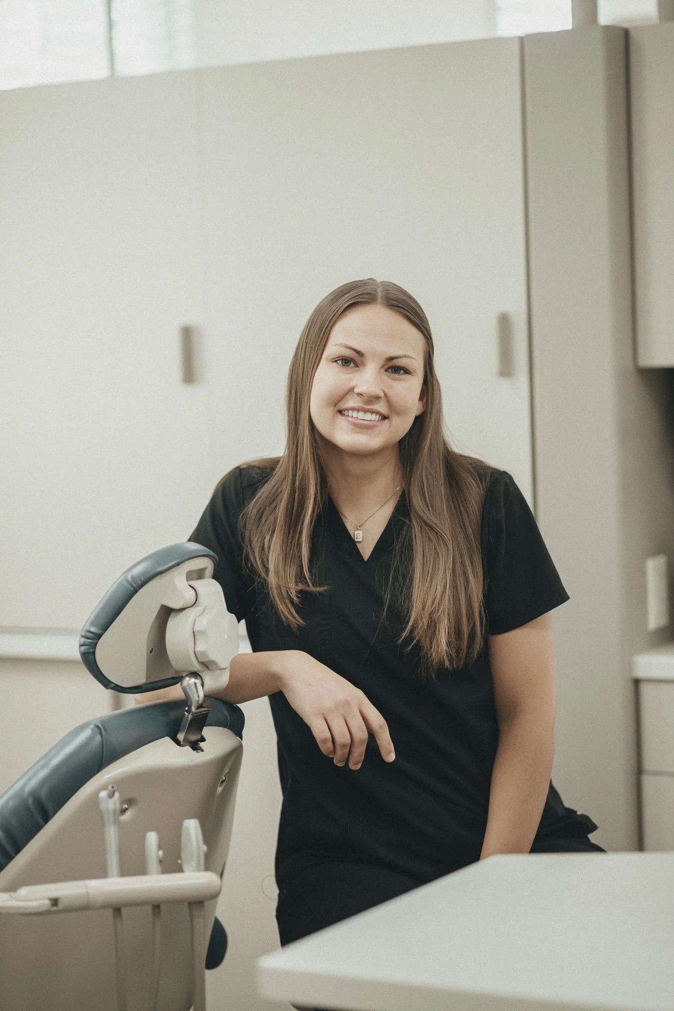 A woman with long brown hair and a black uniform sitting in a dental clinic next to dental equipment.