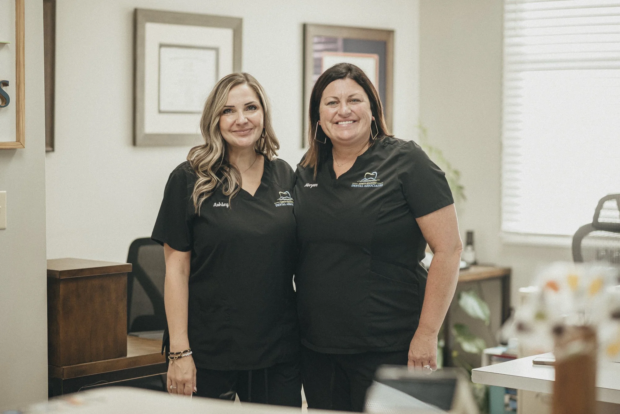 Two women wearing black dental uniforms standing in an office with framed artwork on the wall behind them and a window on the right, smiling at the camera.