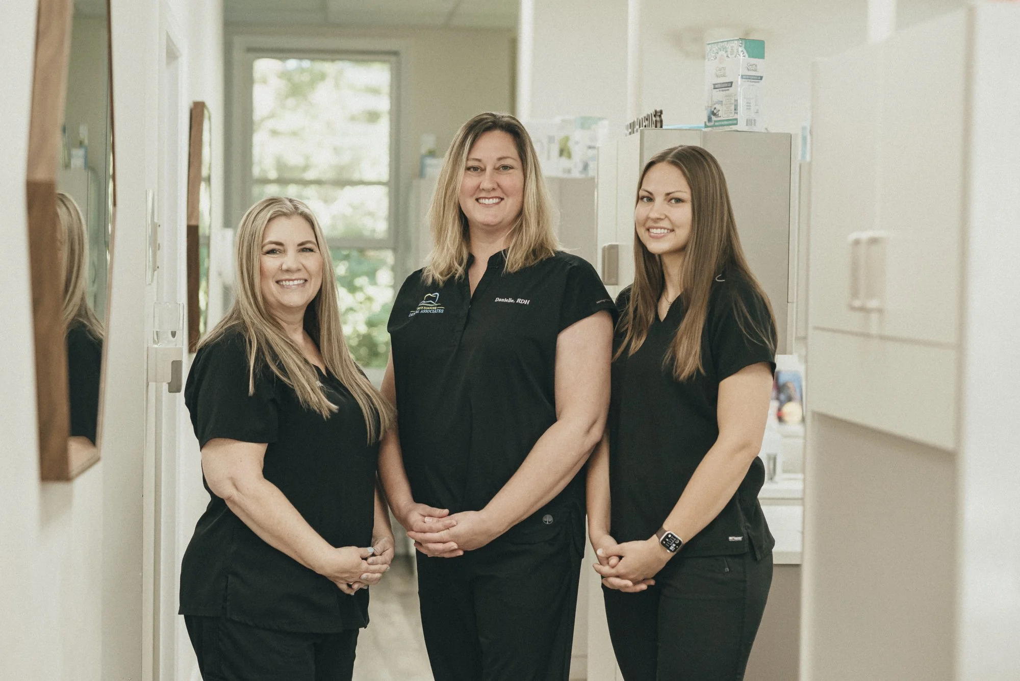 Three women wearing black scrubs standing in a medical or dental clinic hallway, smiling at the camera.