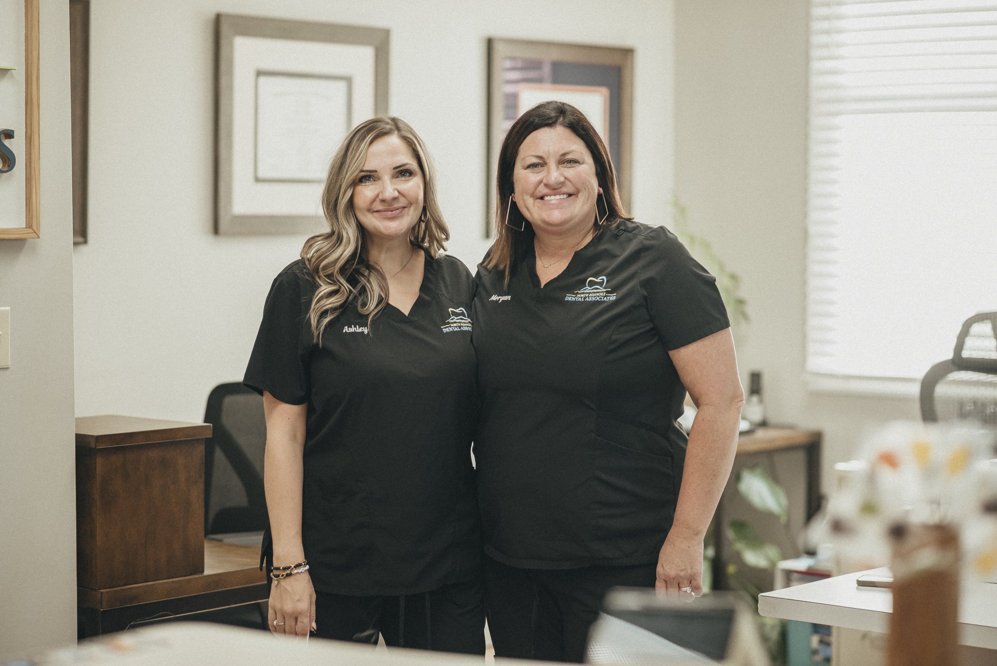 Two women in black scrubs standing in an office, smiling at the camera.