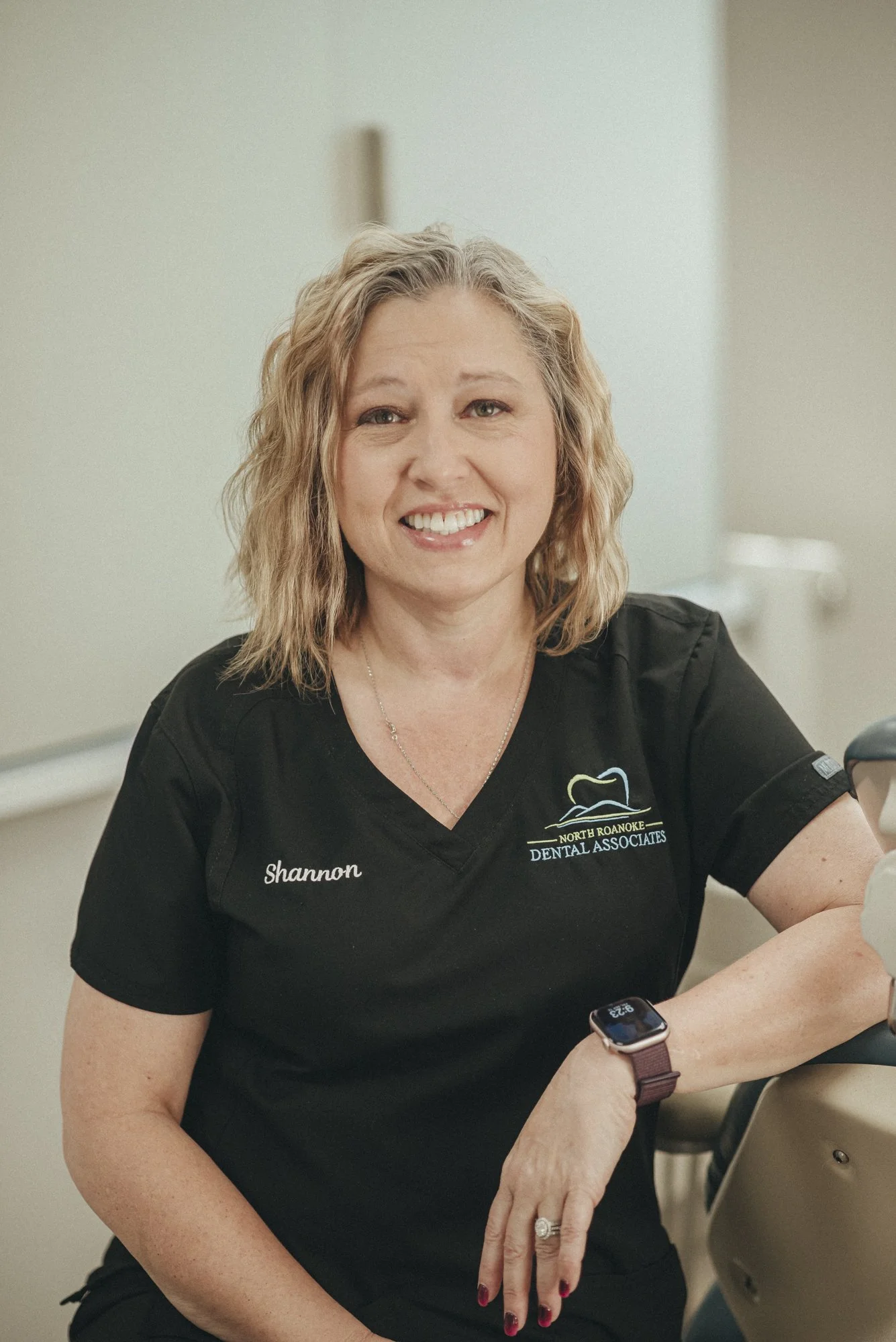A woman with shoulder-length blonde hair, wearing a black dental uniform with the name 'Shannon' embroidered, smiling at the camera inside a dental office.