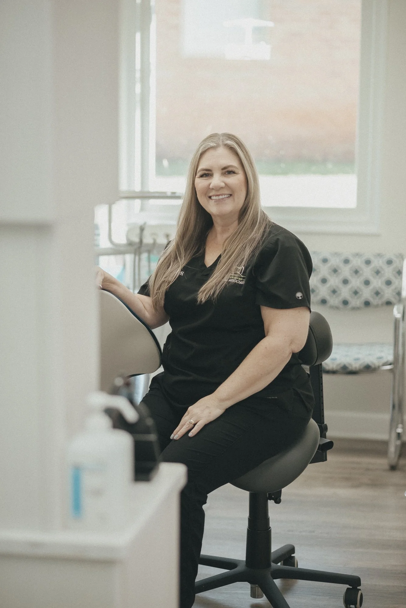 A smiling woman in medical scrubs seated on a rolling stool in a clinical setting, with a window and medical equipment in the background.