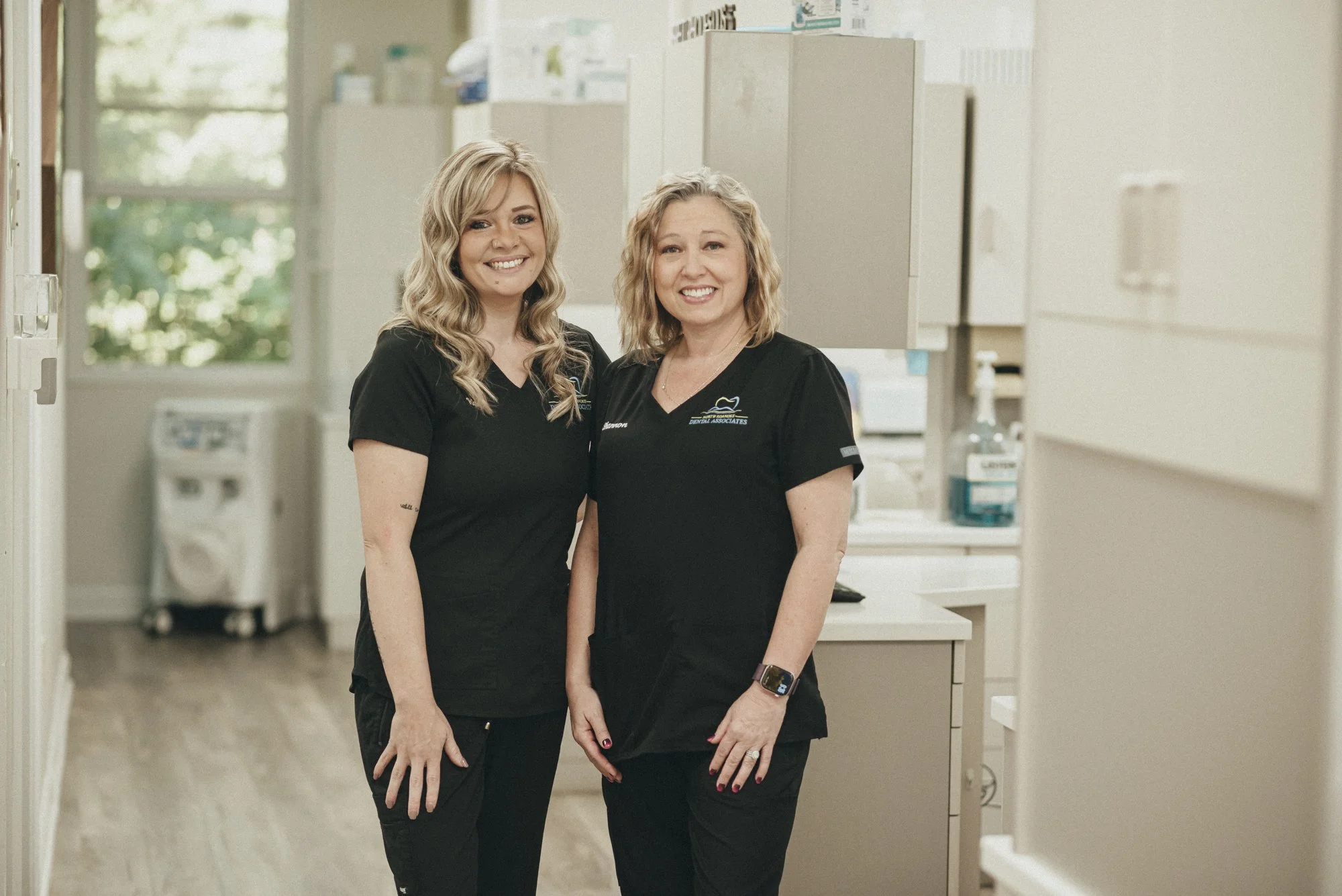 Two women wearing black scrubs standing in a medical or dental office, smiling at the camera.