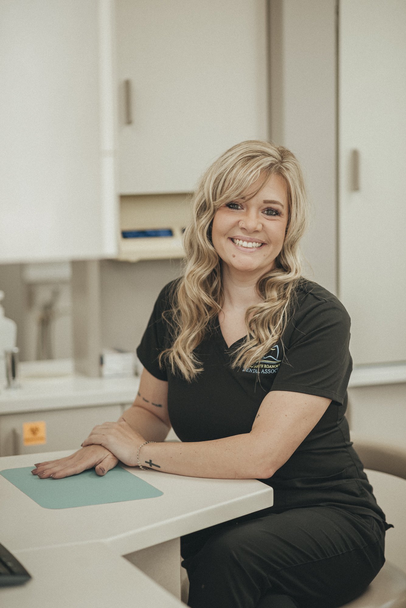 A woman with long blonde curly hair smiling while sitting at a dental office reception desk.