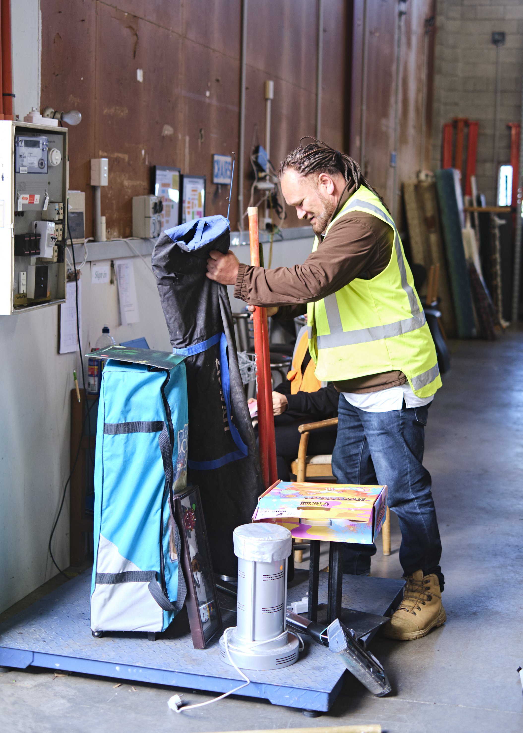A man in a yellow safety vest is standing in a warehouse, climbing on a scale, with various items around him, including a large blue and black bag, a box of toys, and a white humidifier, with a woman seated in the background and industrial equipment on the wall.