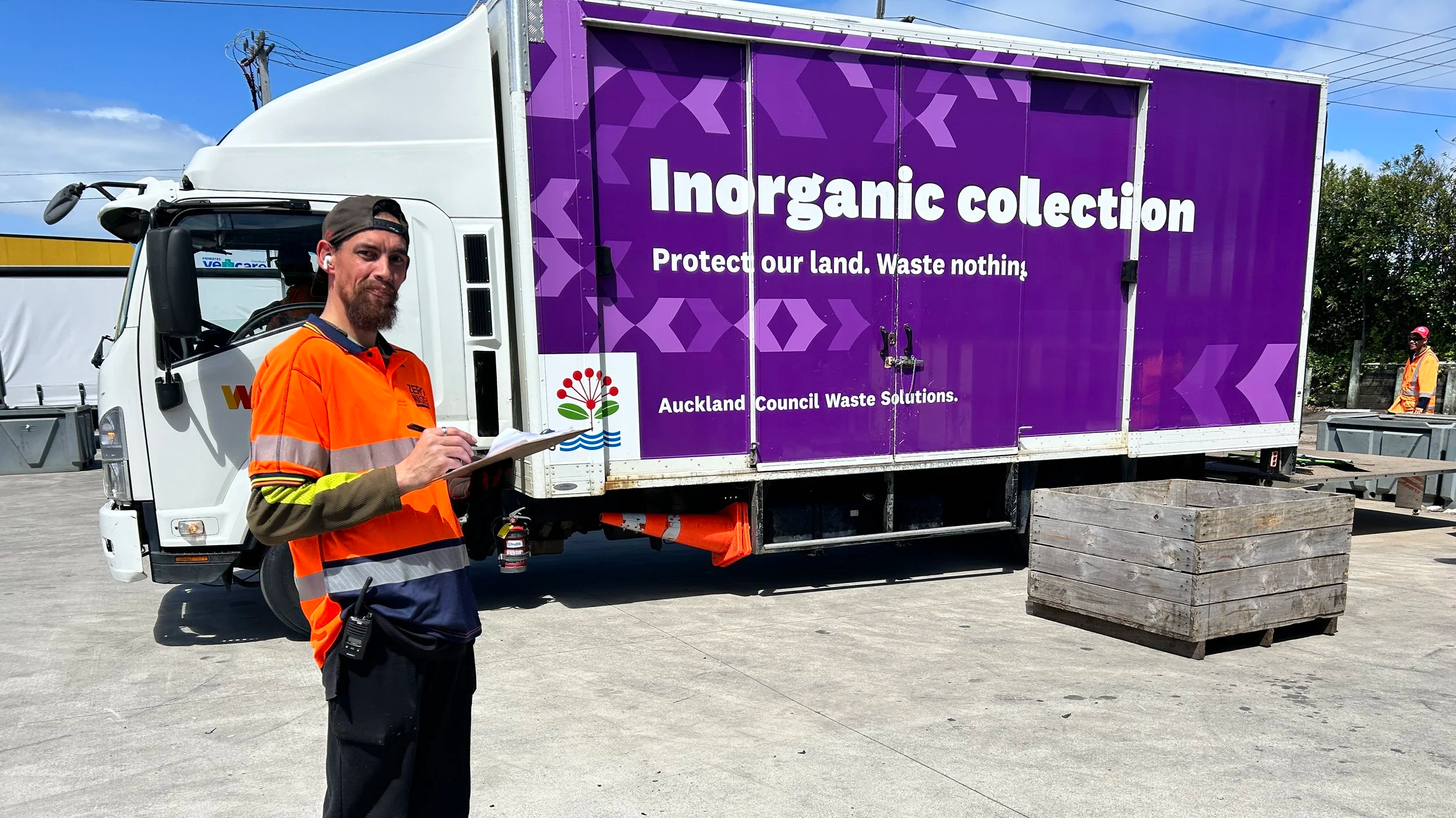A man in an orange and yellow safety vest writes on a clipboard in front of a truck with purple signage promoting Inorganic Collections for Auckland Council Waste Solutions. Two other workers are visible in the background.