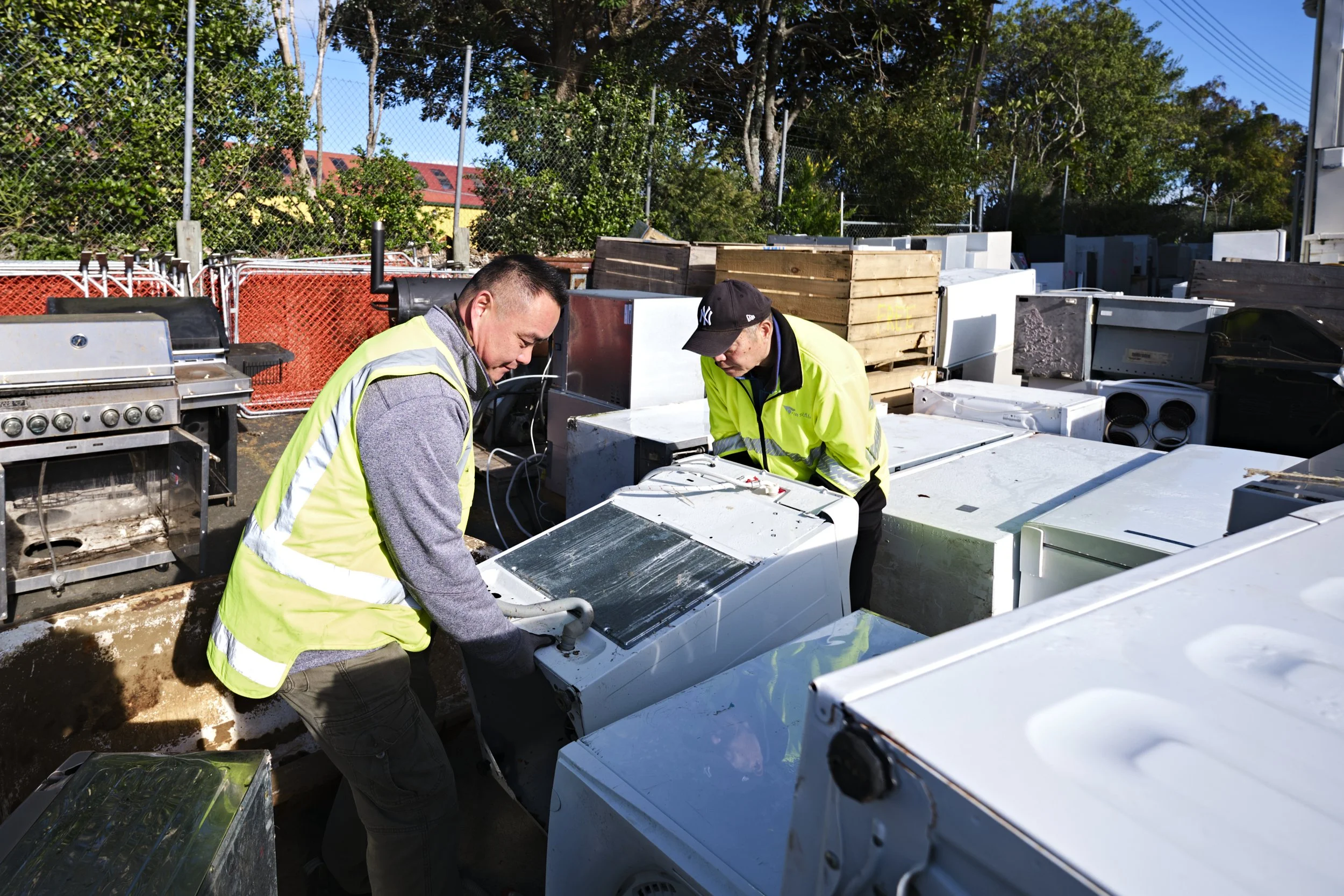 Two workers in neon safety vests examining washing machine appliances outdoors with various whiteware and appliances around, behind a chain-link fence and trees under a blue sky.