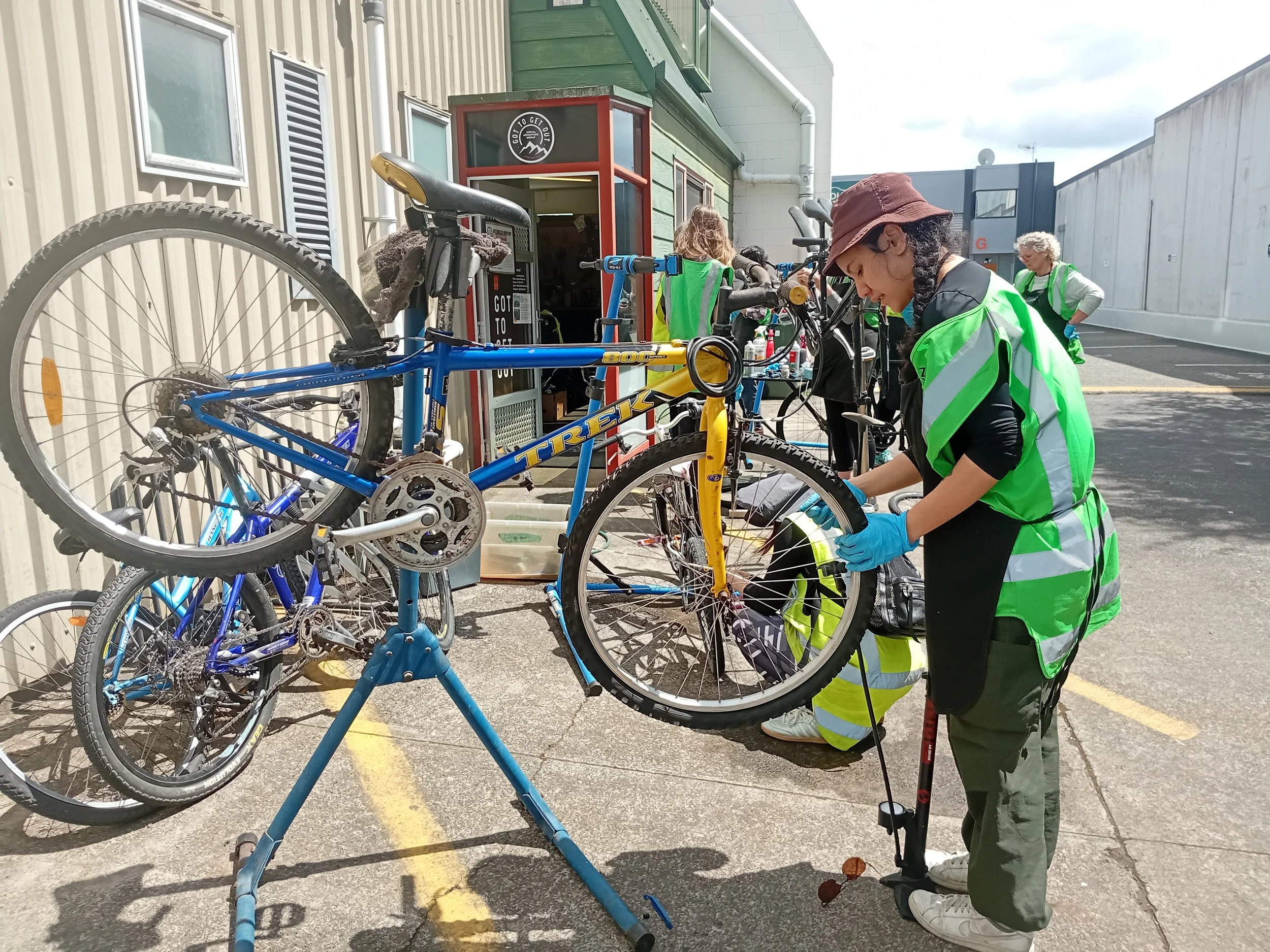 A woman in a green safety vest and gloves repairing a bicycle at a bike repair station outside a building, with other cyclists nearby.