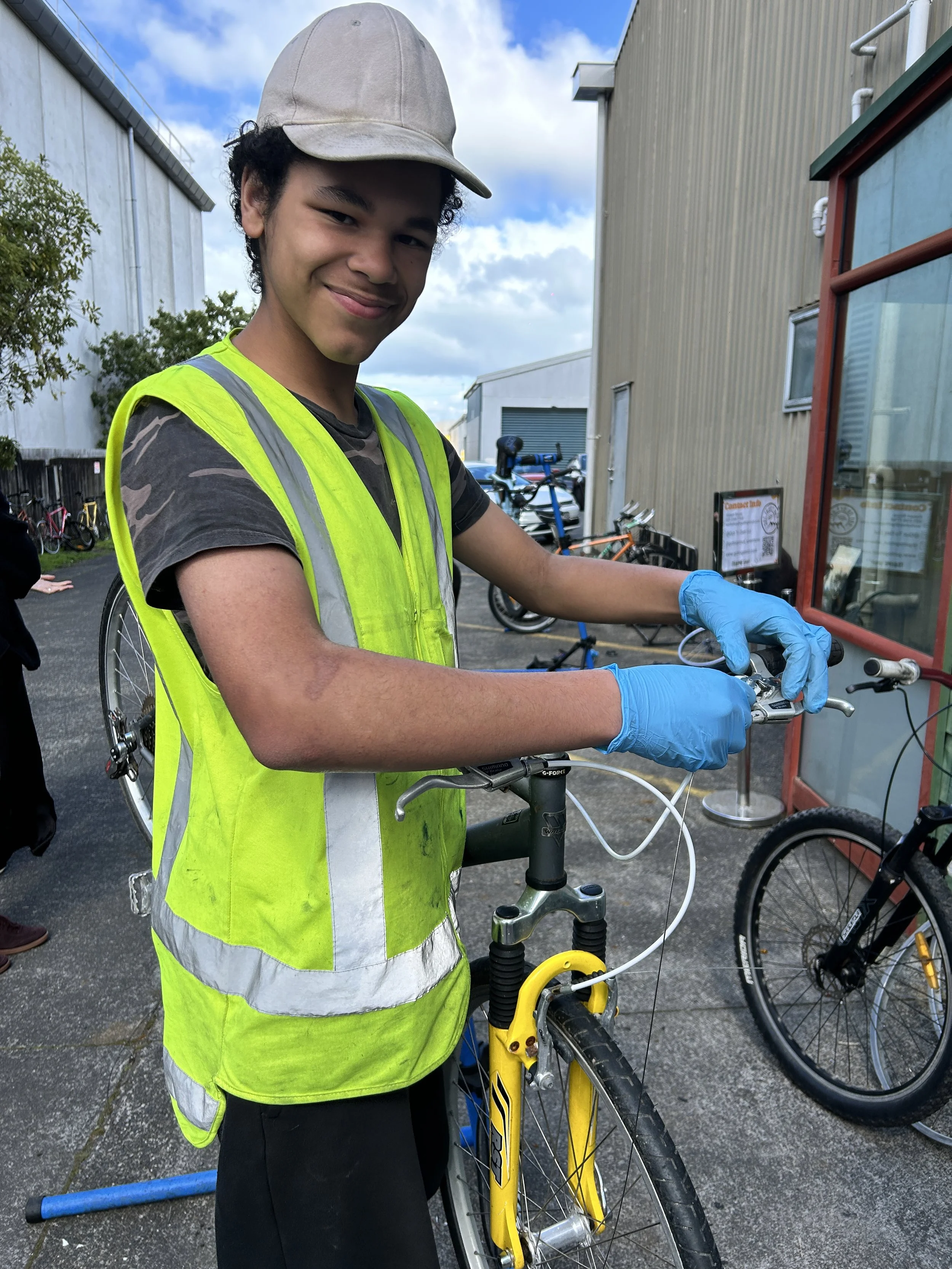 A young man with curly hair wearing a beige cap, neon yellow safety vest, and blue gloves, standing outside and fixing a yellow bicycle.