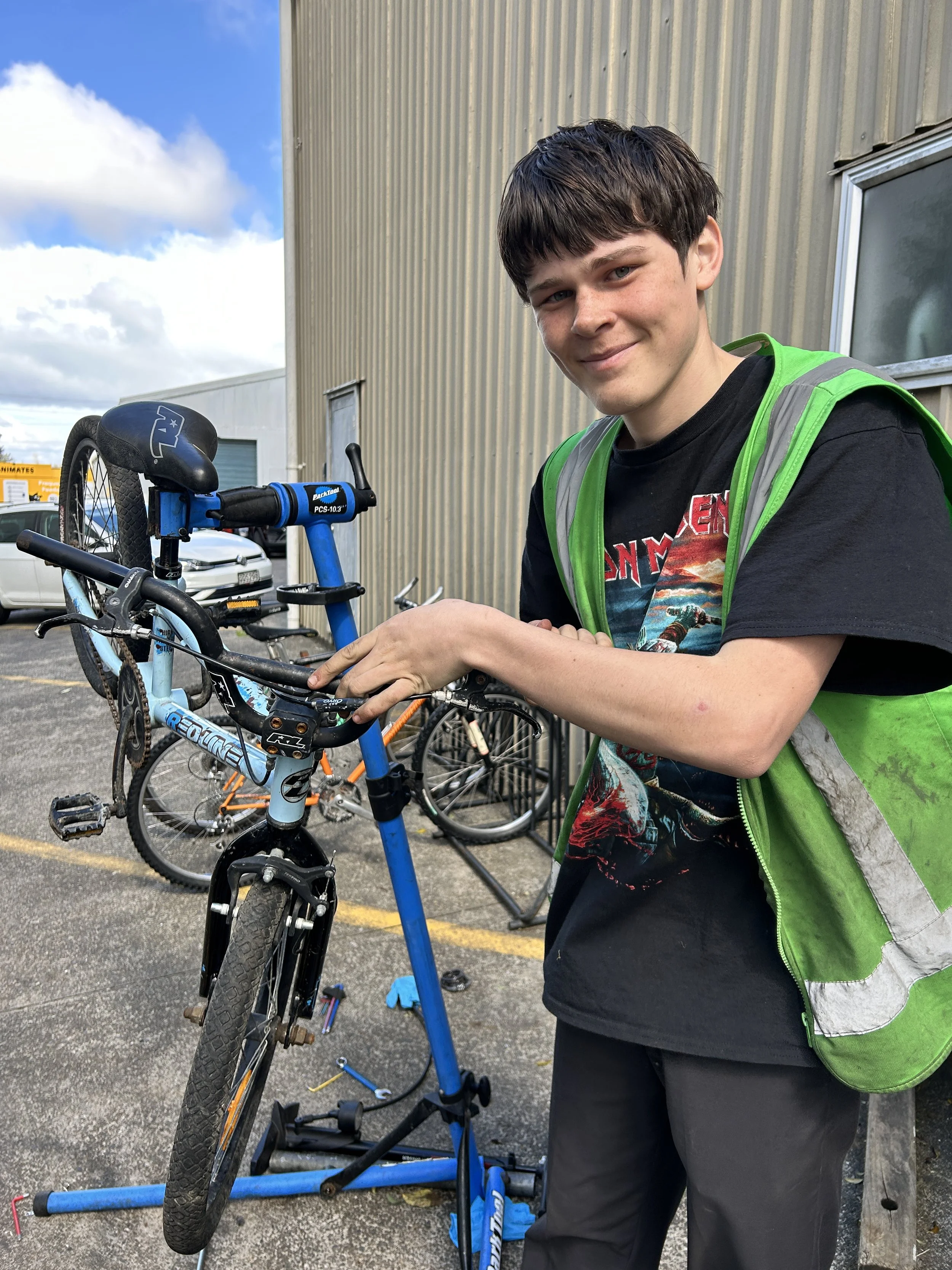 A young man with dark hair wearing a black Iron Maiden t-shirt and a green safety vest working on a blue mountain bike outside a building with a parking lot and other bikes behind him.