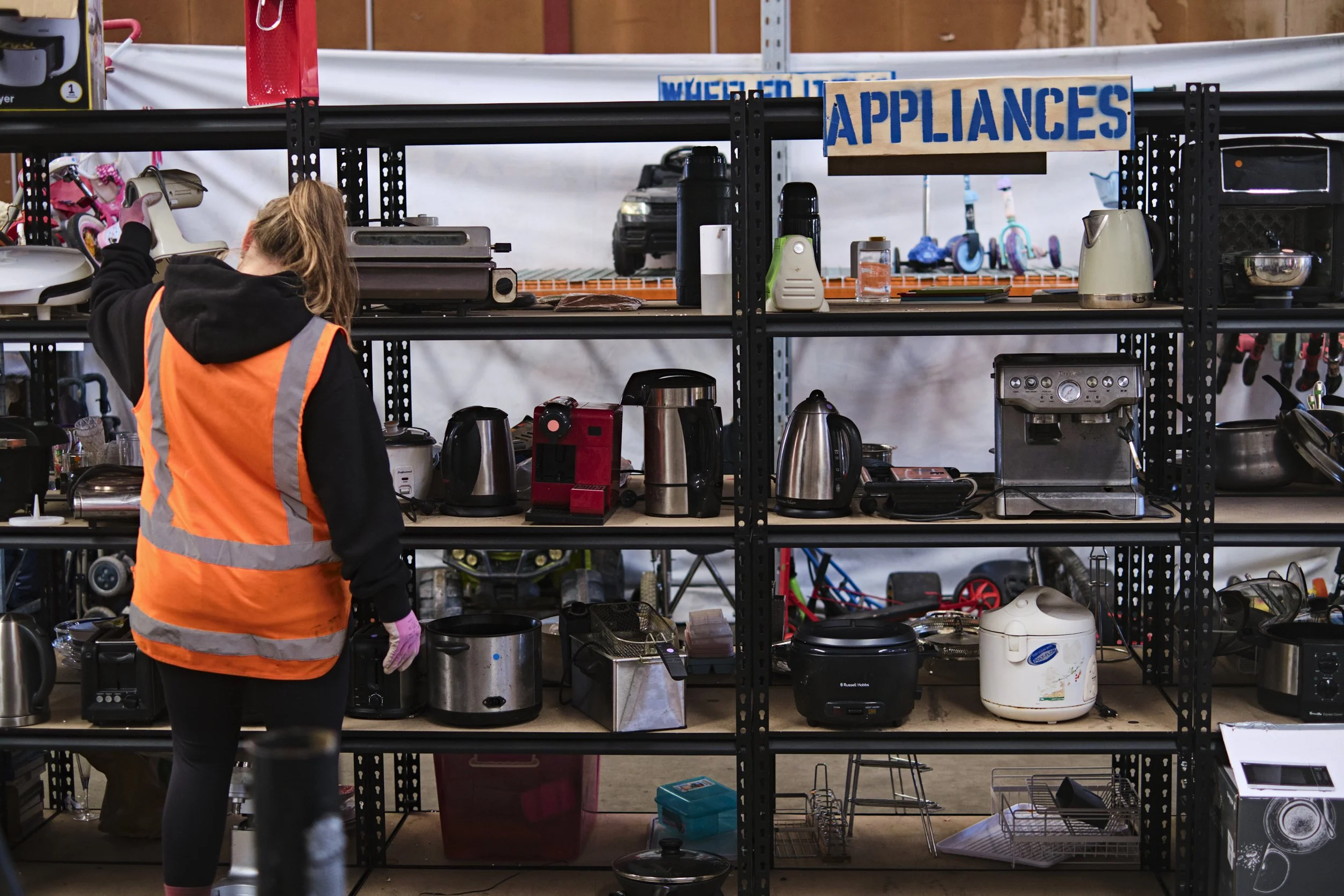 A woman wearing an orange safety vest and black hoodie is shopping for appliances at an op shop, browsing shelves filled with kitchen appliances and second hand items.