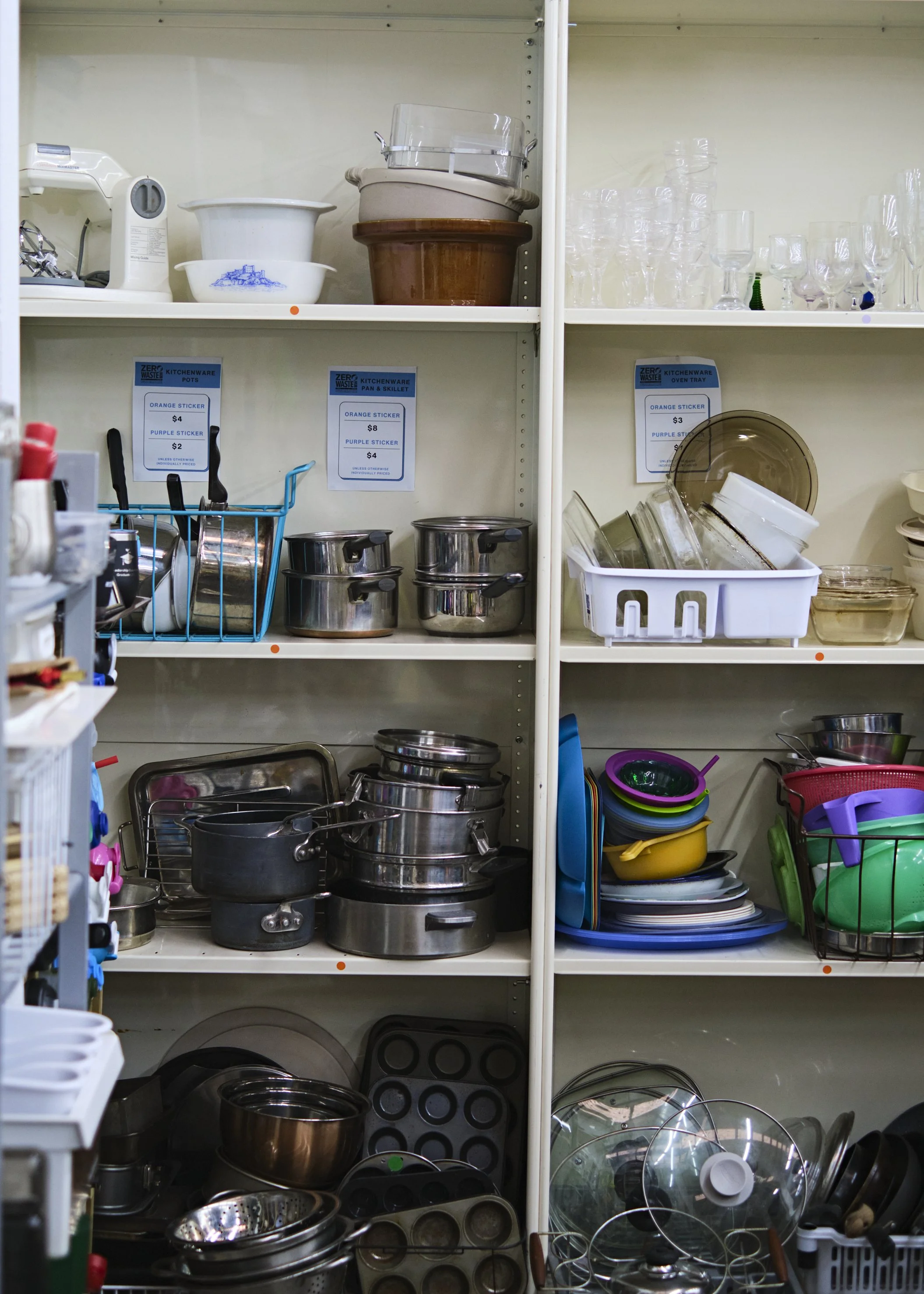 Shelf filled with various kitchenware including pots, pans, glasses, bowls, and containers in a thrift store or kitchen supply store.